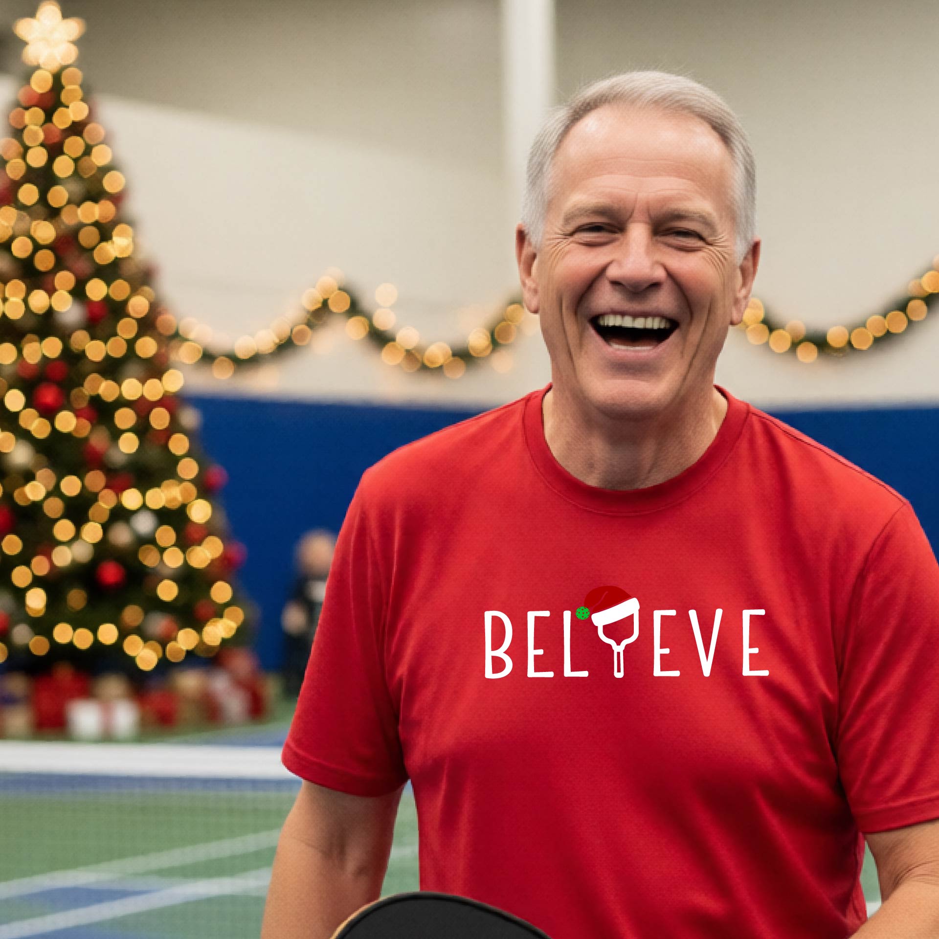 A man wearing a red short-sleeve t-shirt with a  pickleball graphic with text "BELIEVE" featuring a the letter I replaced with a pickleball paddle wearing a Santa hat.  