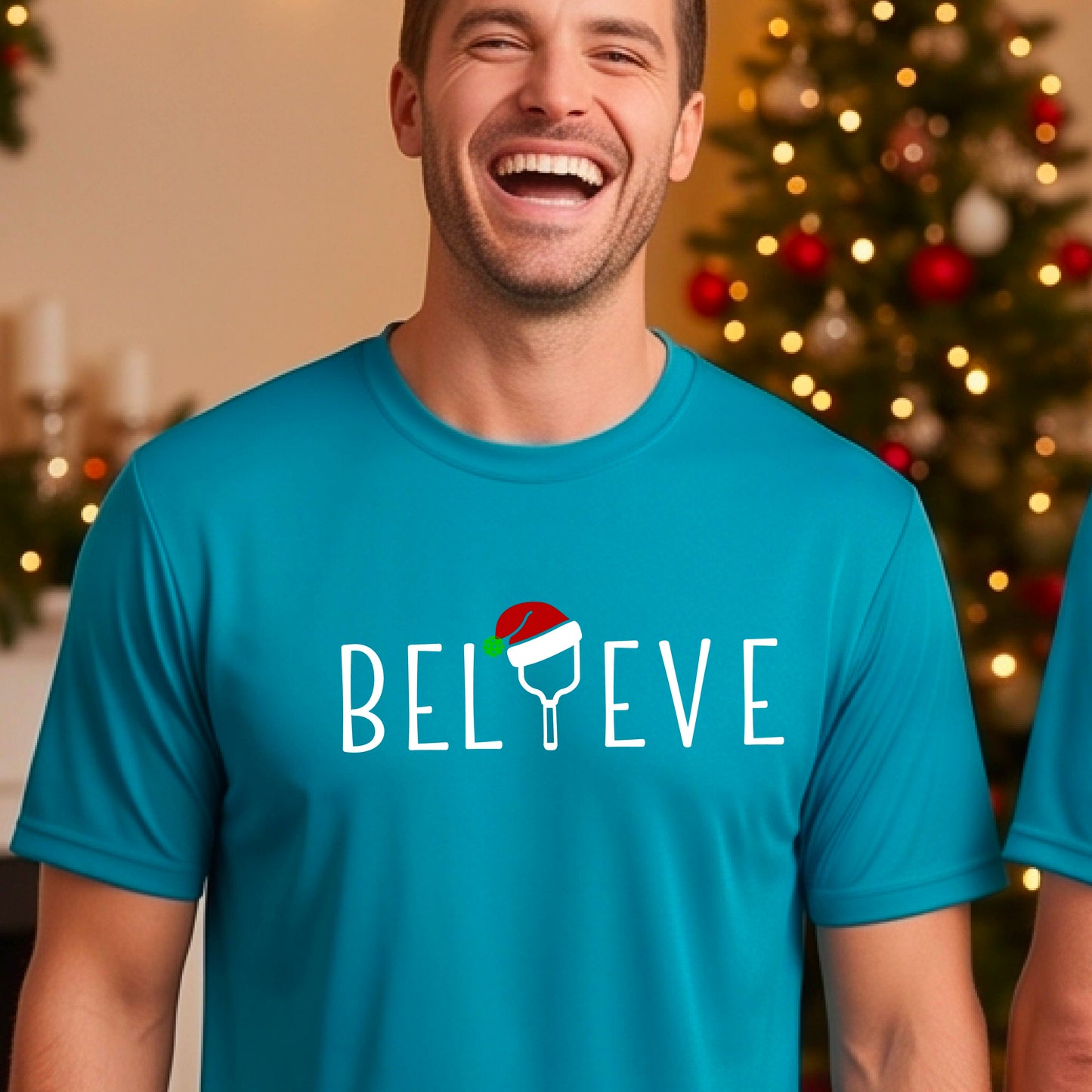 A man wearing a bright blue short-sleeve t-shirt with a  pickleball graphic with text "BELIEVE" featuring a the letter I replaced with a pickleball paddle wearing a Santa hat.  