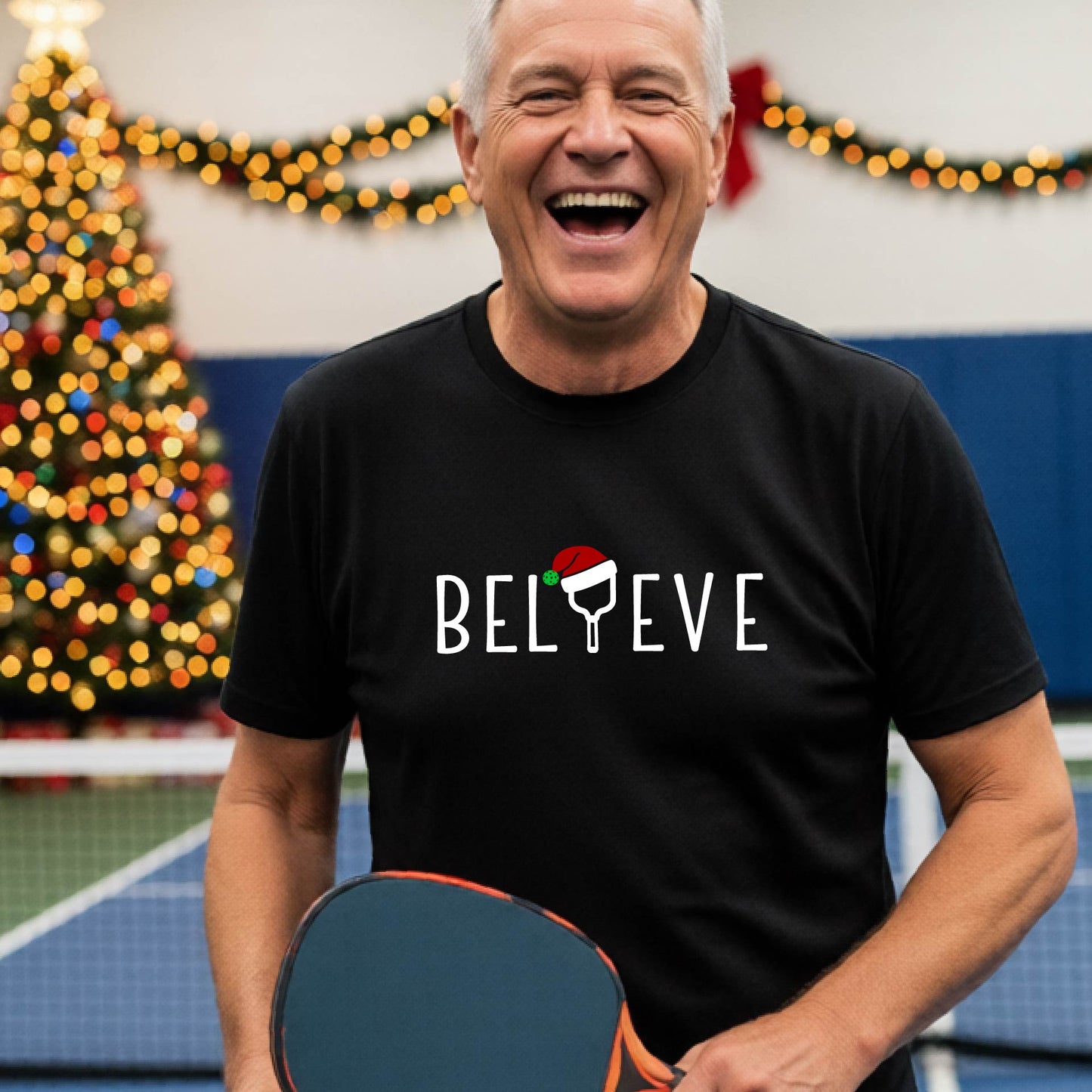 A man wearing an black short-sleeve t-shirt with a  pickleball graphic with text "BELIEVE" featuring a the letter I replaced with a pickleball paddle wearing a Santa hat.  