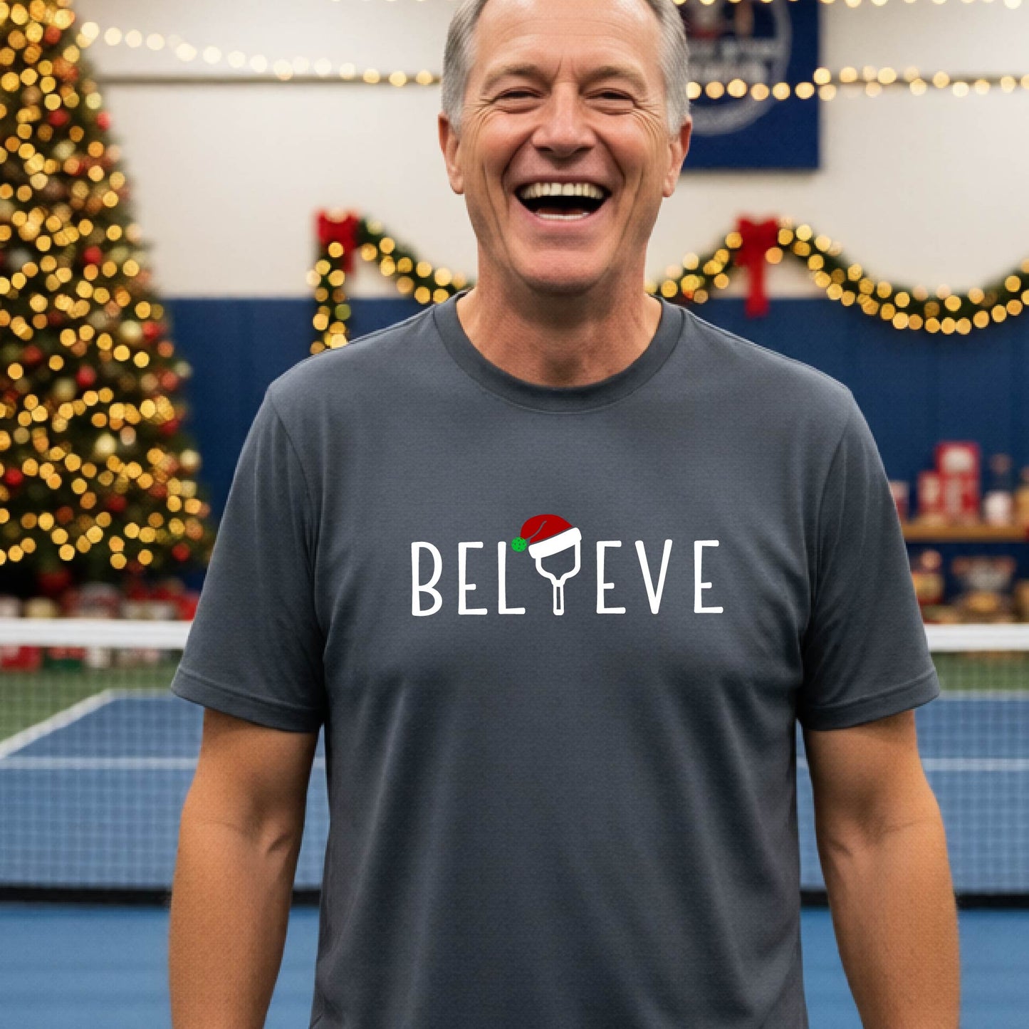 A man wearing a gray short-sleeve t-shirt with a  pickleball graphic with text "BELIEVE" featuring a the letter I replaced with a pickleball paddle wearing a Santa hat.  