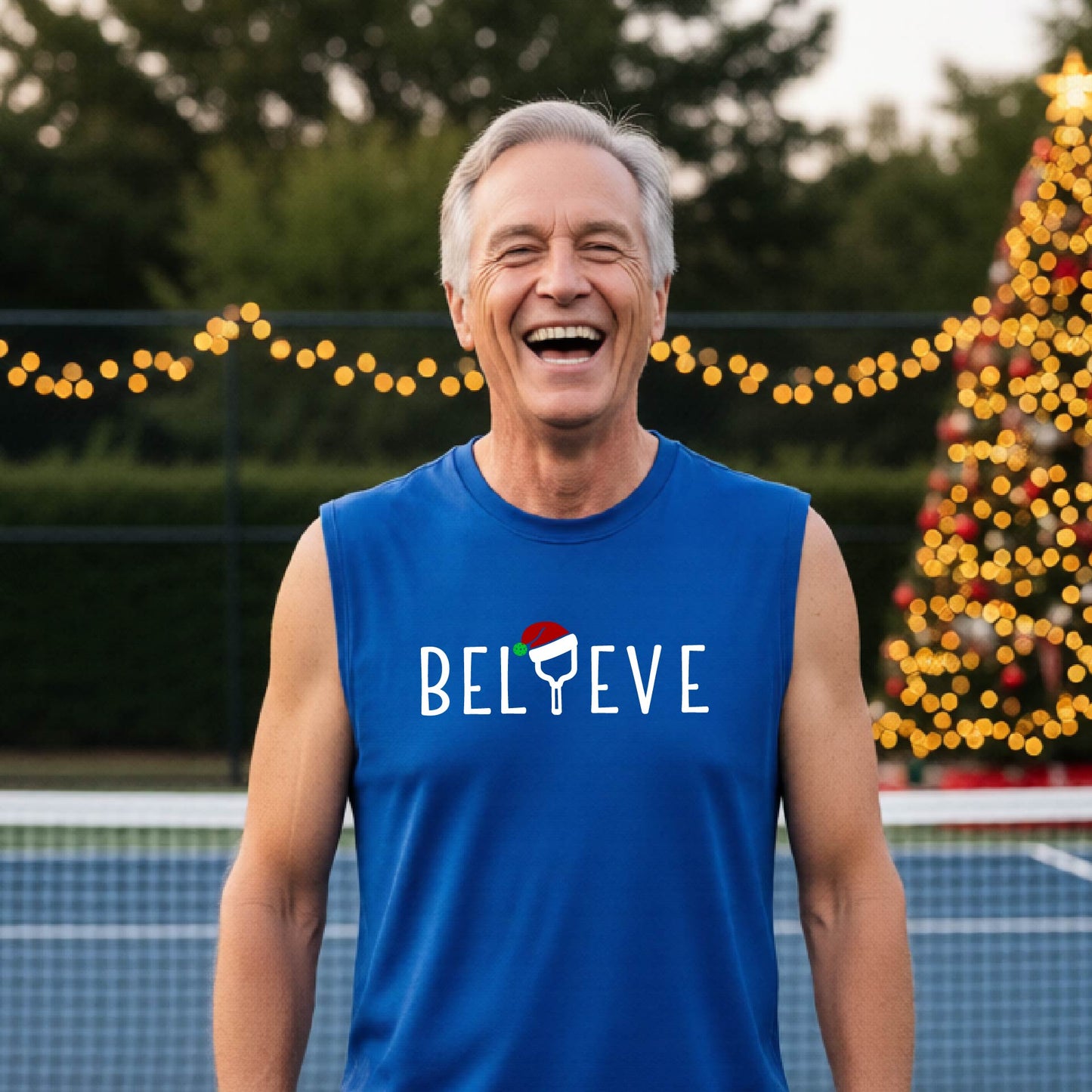 A smiling man wearing a royal blue sleeveless shirt with the word 'BELIEVE' with the I repalced with a pickleball paddle wearing a Santa hat graphic stands on a pickleball court with a Christmas tree iand lights n the background.