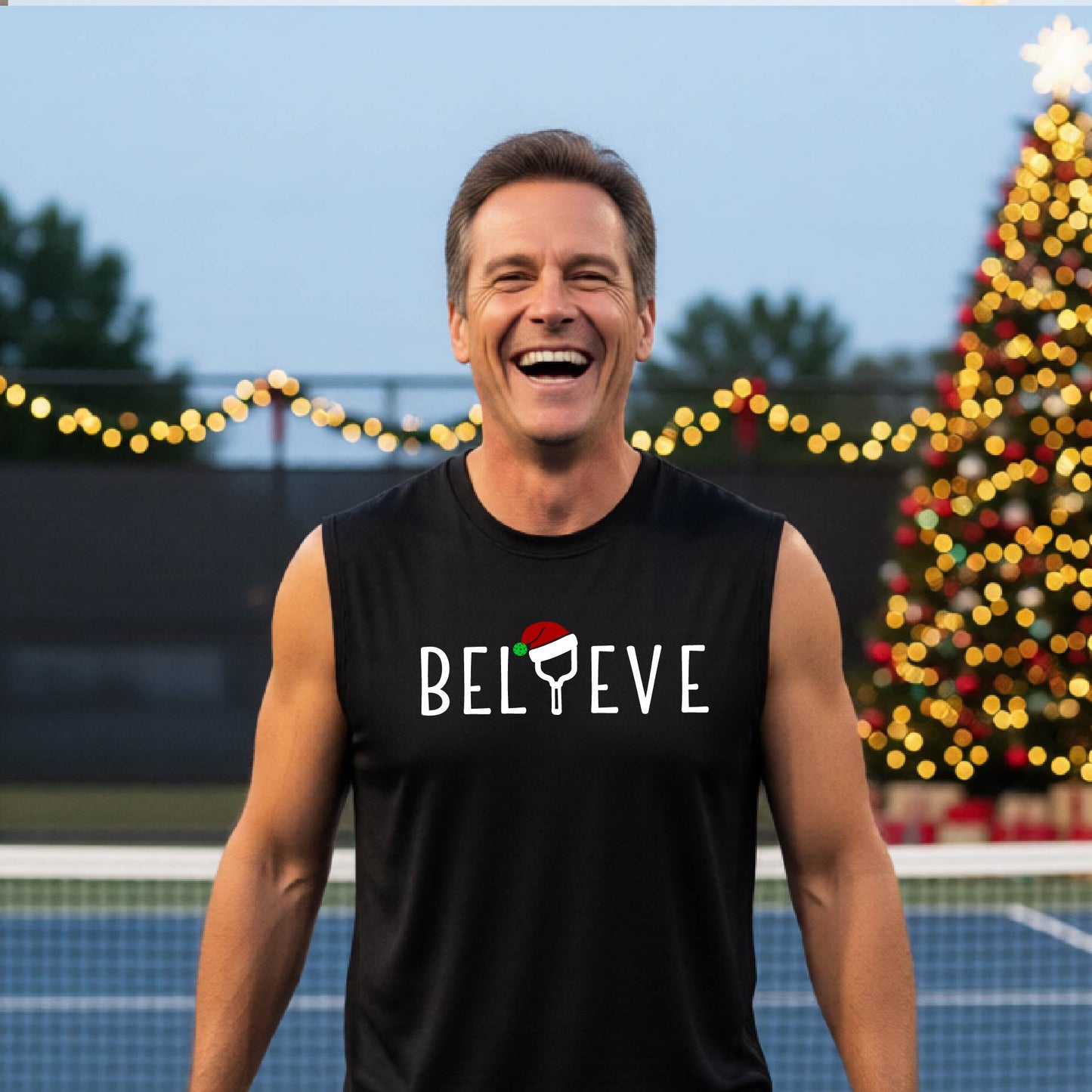 A smiling man wearing a black sleeveless shirt with the word 'BELIEVE' with the I repalced with a pickleball paddle wearing a Santa hat graphic stands on a pickleball court with a Christmas tree iand lights n the background.