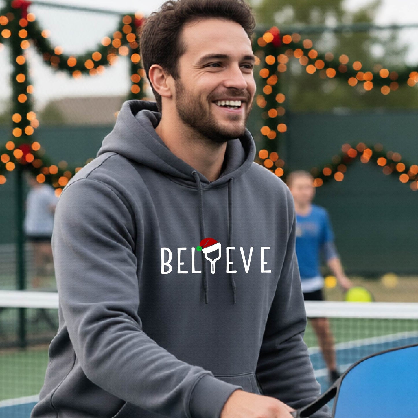 A man wearing a gray hoodie with a white 'BELIEVE' graphic with a pickleball paddle wearing a Santa hat in front of a pickleball court. 