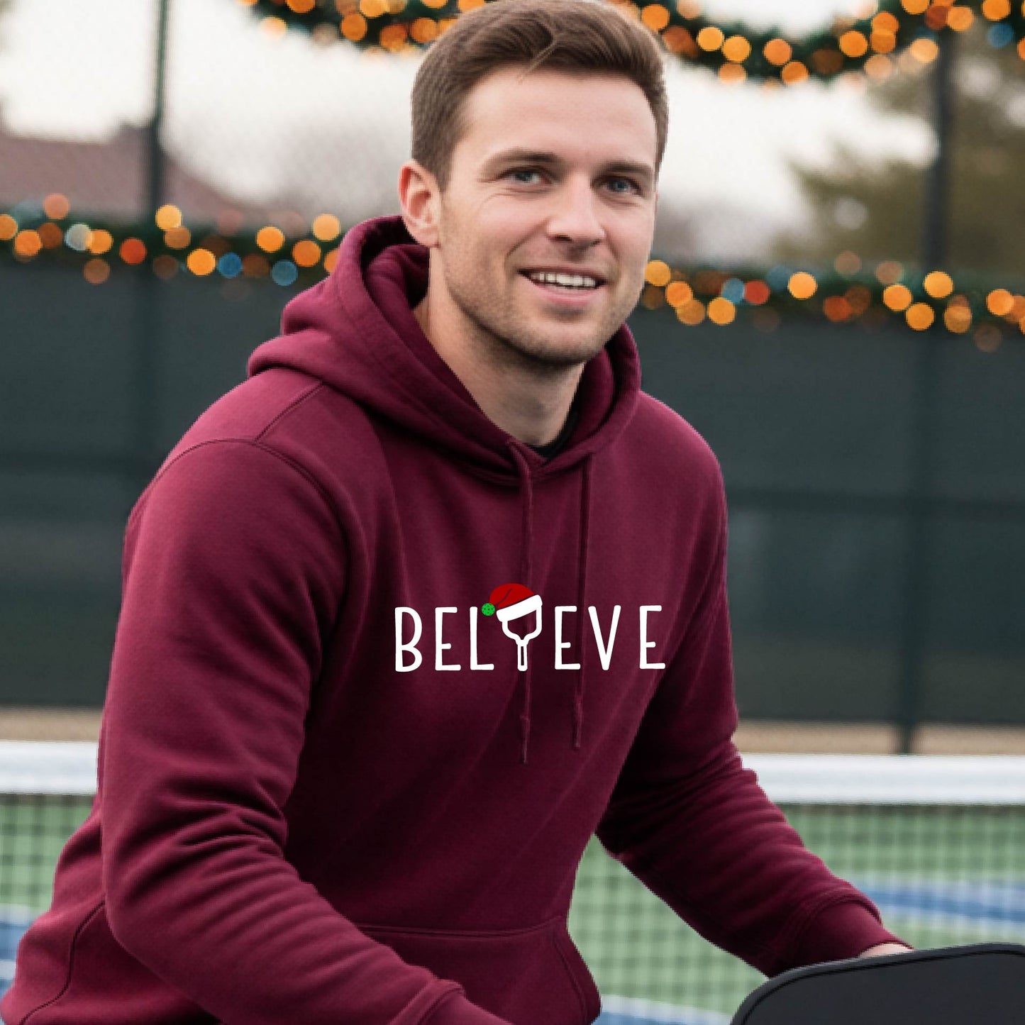 A man wearing a maroon hoodie with a white 'BELIEVE' graphic with a pickleball paddle wearing a Santa hat in front of a pickleball court. 