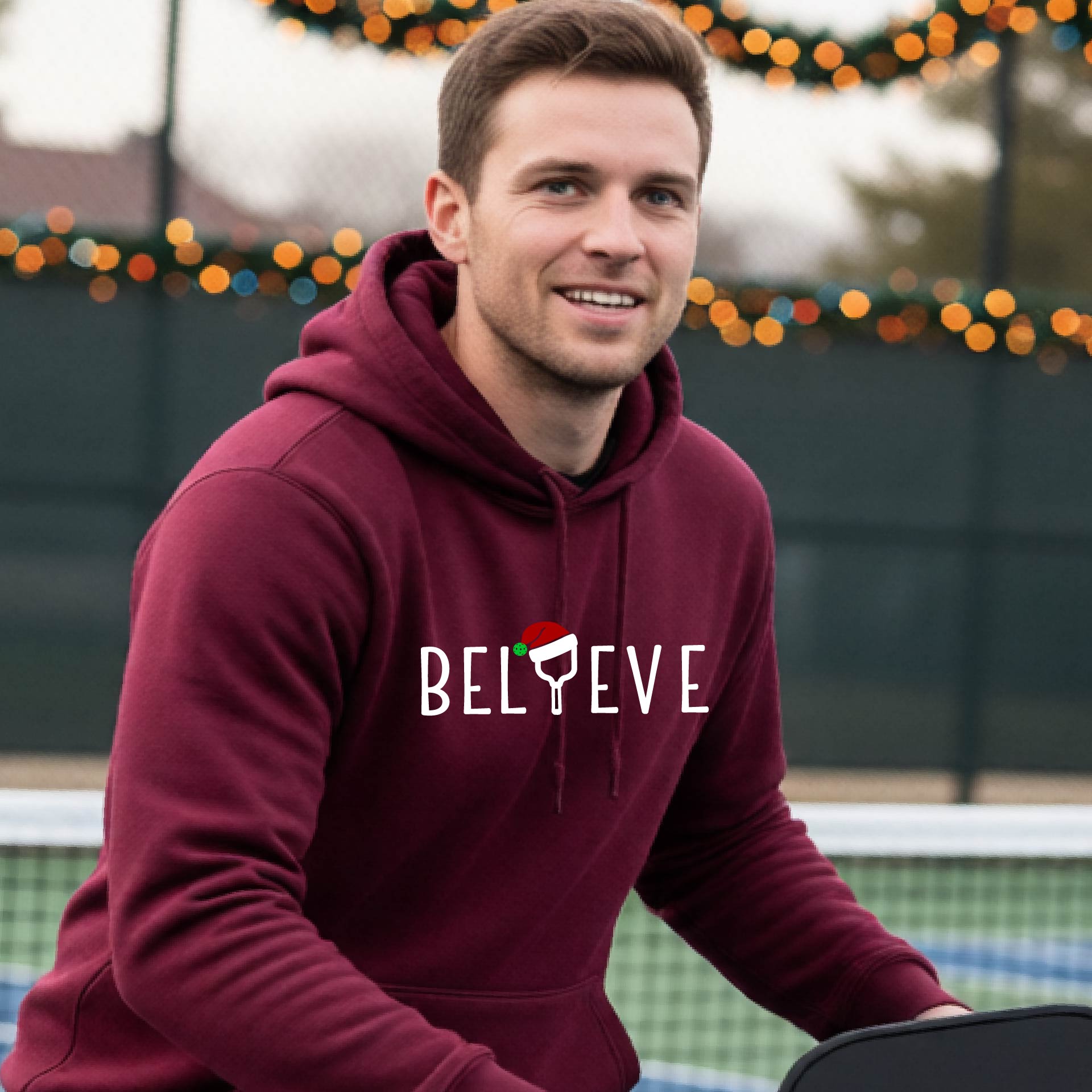 A man wearing a maroon hoodie with a white 'BELIEVE' graphic with a pickleball paddle wearing a Santa hat in front of a pickleball court. 