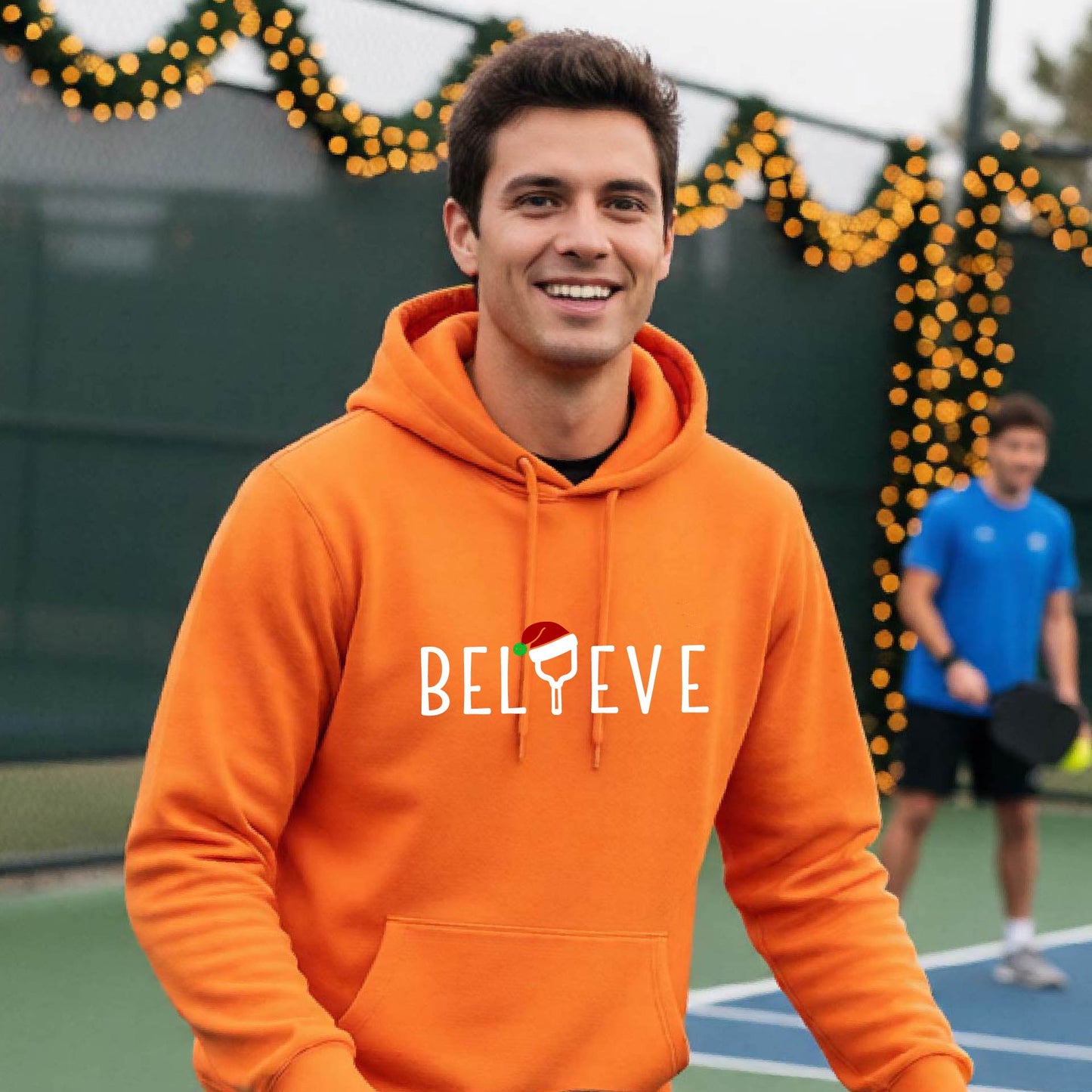 A man wearing a bright orange  hoodie with a white 'BELIEVE' graphic with a pickleball paddle wearing a Santa hat in front of a pickleball court. 