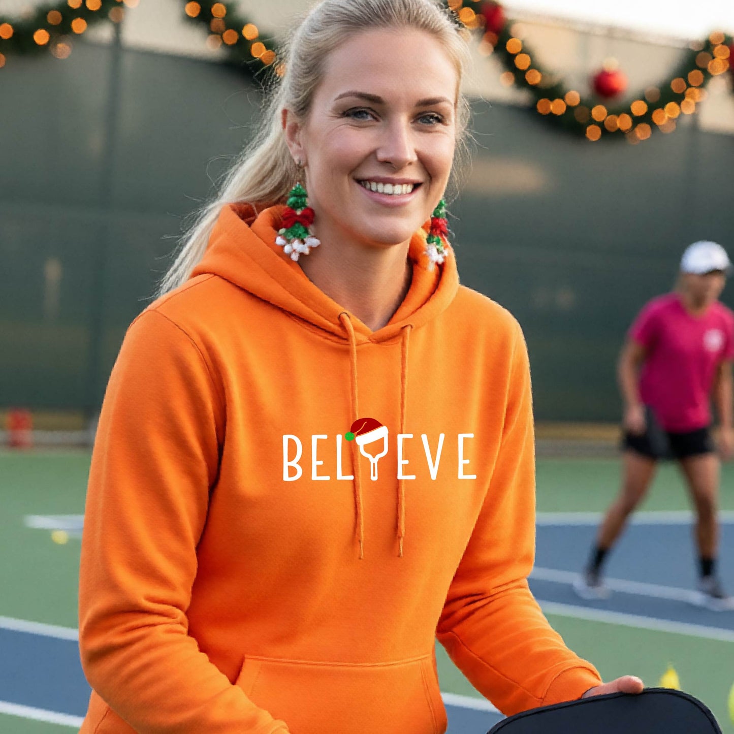 A woman wearing an orange hoodie with a white 'BELIEVE' graphic with a pickleball paddle wearing a Santa hat in front of a pickleball court. 