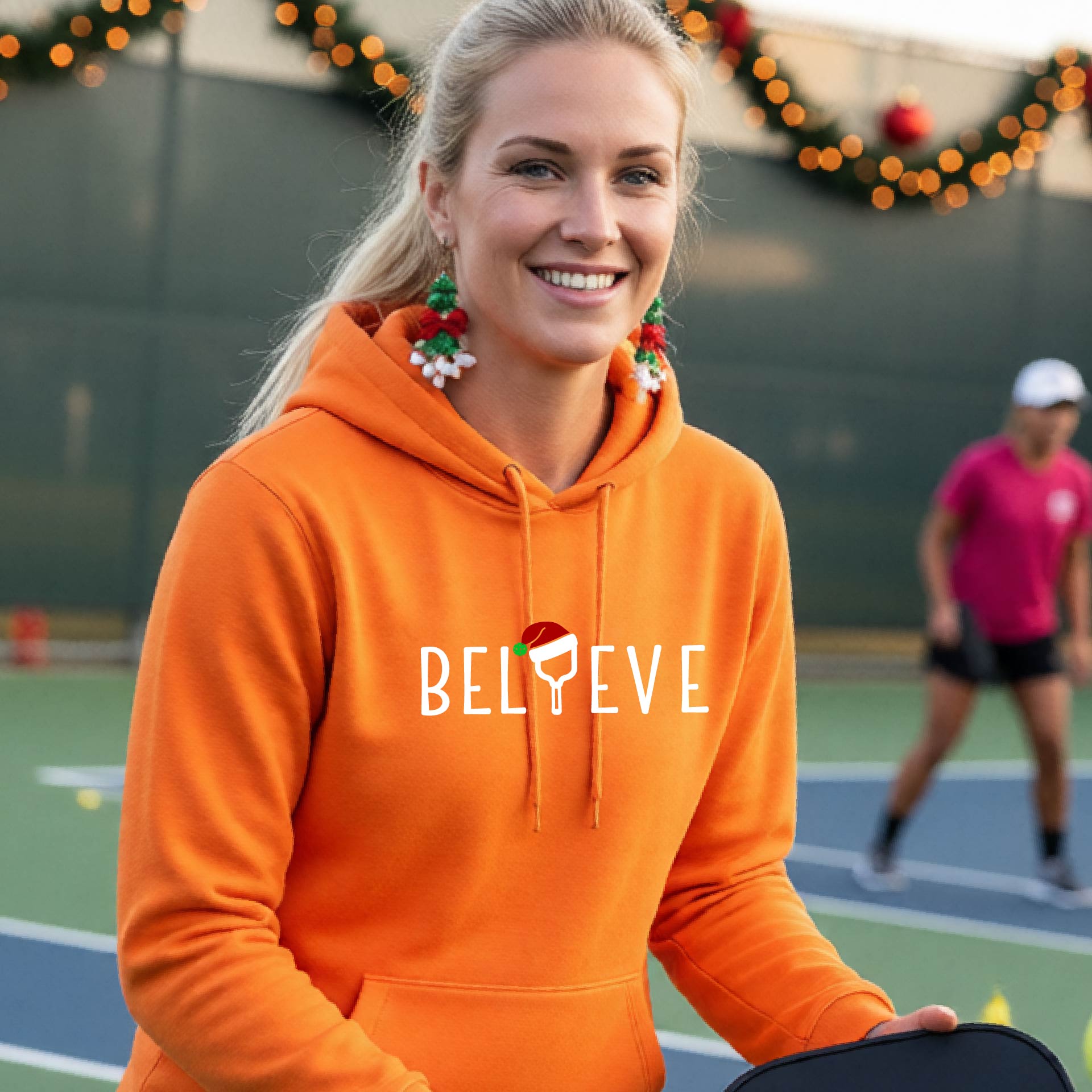 A woman wearing an orange hoodie with a white 'BELIEVE' graphic with a pickleball paddle wearing a Santa hat in front of a pickleball court. 