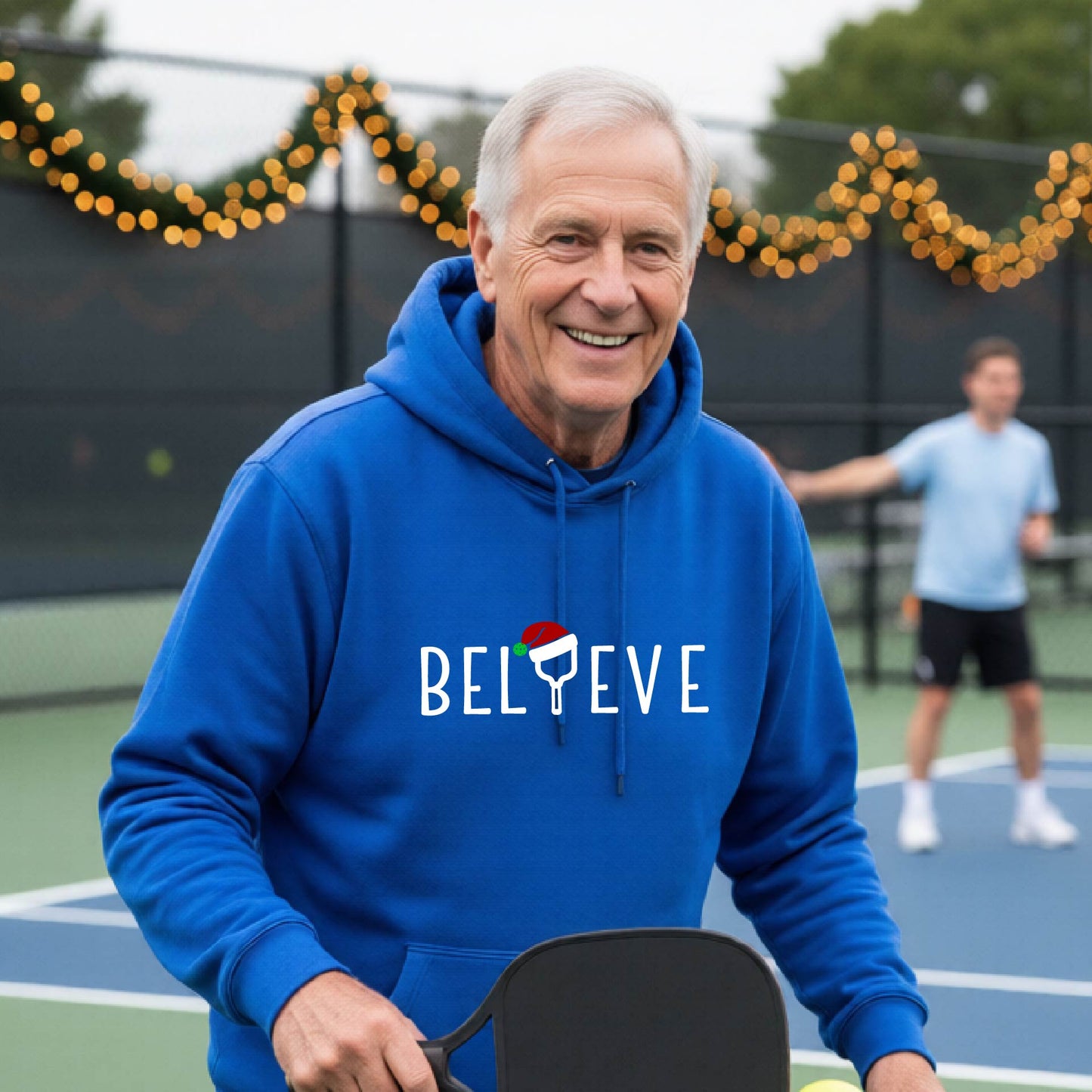 A man wearing a royal blue hoodie with a white 'BELIEVE' graphic with a pickleball paddle wearing a Santa hat in front of a pickleball court. 
