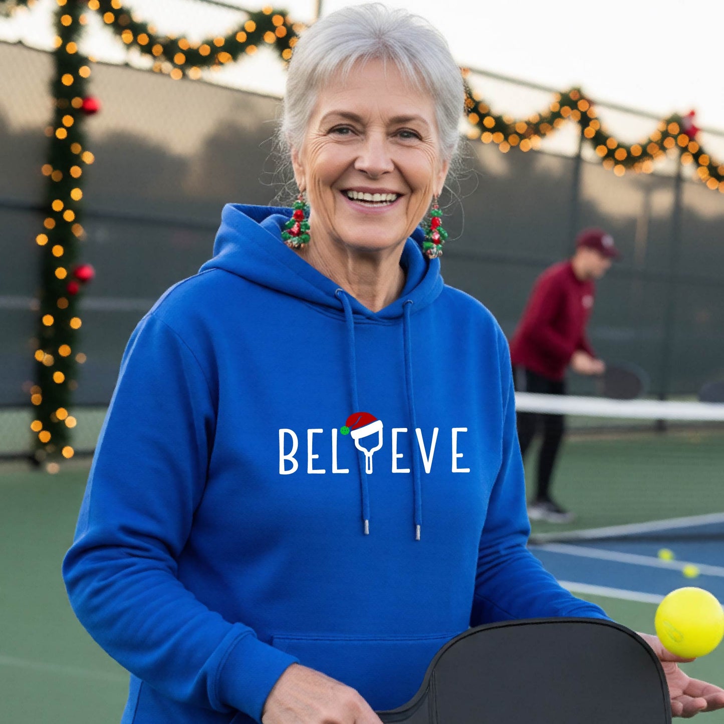 A man wearing a royal blue hoodie with a white 'BELIEVE' graphic with a pickleball paddle wearing a Santa hat in front of a pickleball court. 