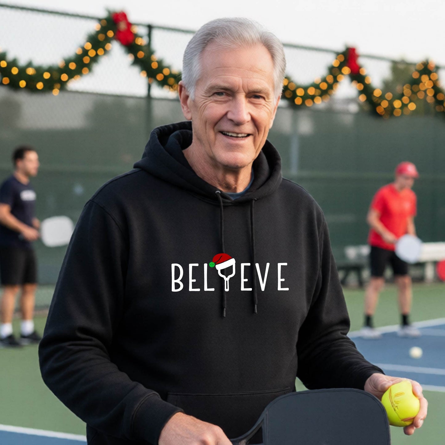 A man wearing a black hoodie with a white 'BELIEVE' graphic with a pickleball paddle wearing a Santa hat in front of a pickleball court. 