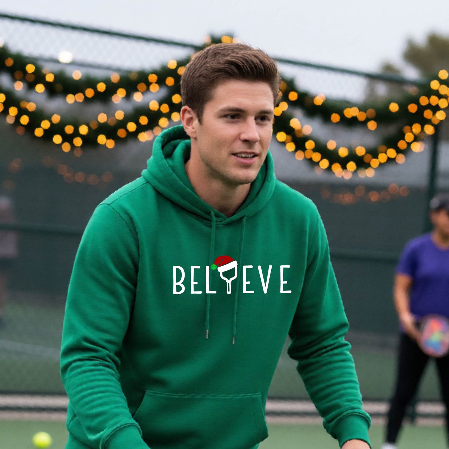 A man wearing a green hoodie with a white 'BELIEVE' graphic with a pickleball paddle wearing a Santa hat in front of a pickleball court. 