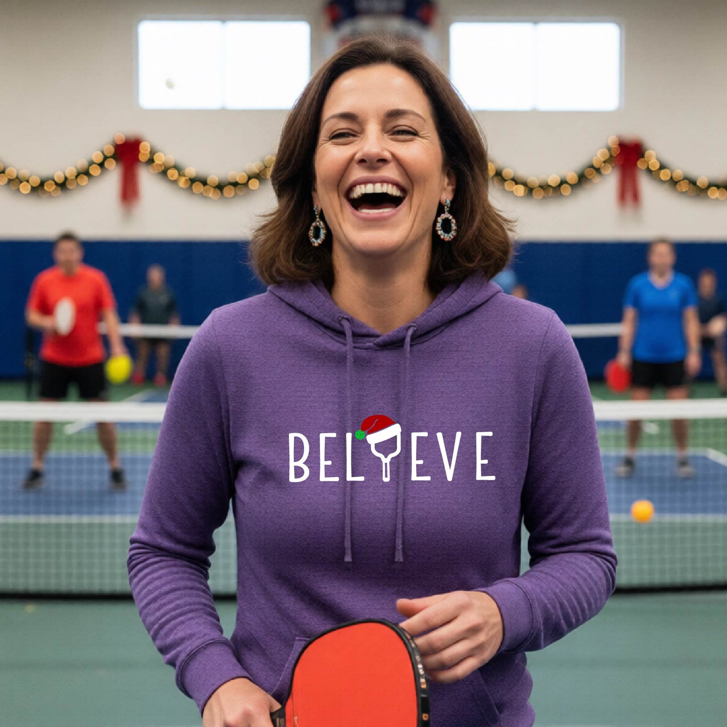 A purple hooded sweatshirt featuring a graphic print that reads "Believe" . The text I is replaced with a pickleball paddle wearing a santa hat.  The backgro0und is Christmas themed. 