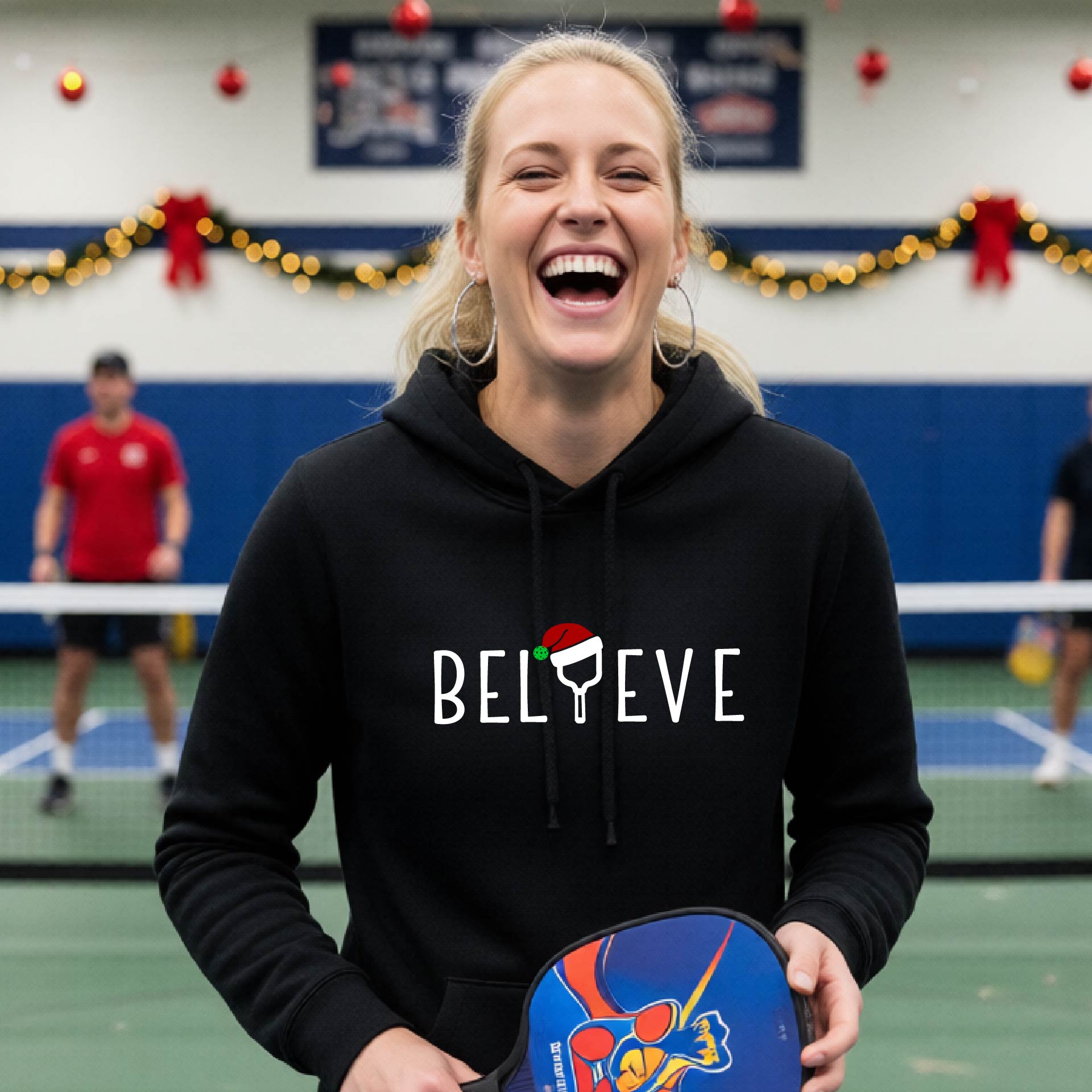 A black hooded sweatshirt featuring a graphic print that reads "Believe" . The text I is replaced with a pickleball paddle wearing a santa hat.  The backgro0und is Christmas themed. 