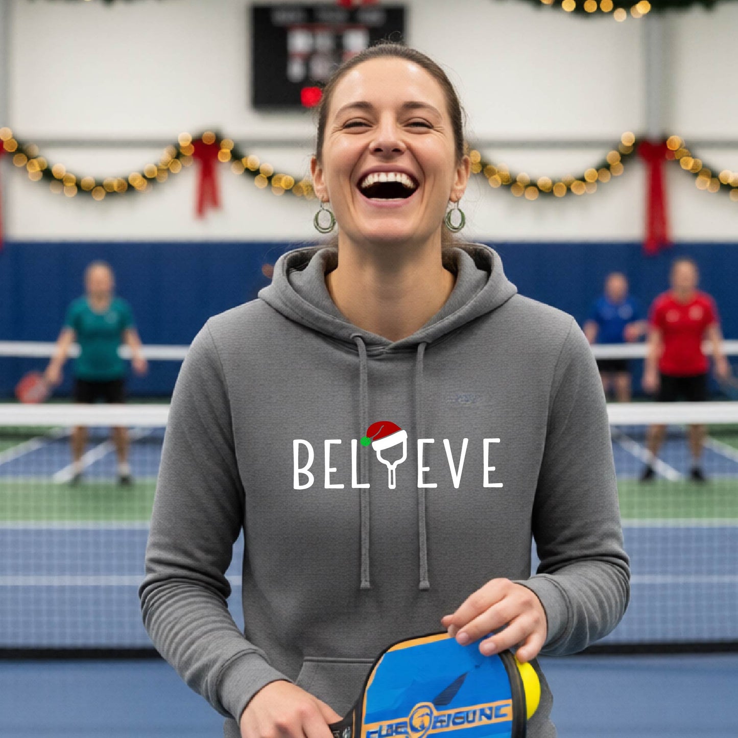 A gray hooded sweatshirt featuring a graphic print that reads "Believe" . The text I is replaced with a pickleball paddle wearing a santa hat.  The backgro0und is Christmas themed. 