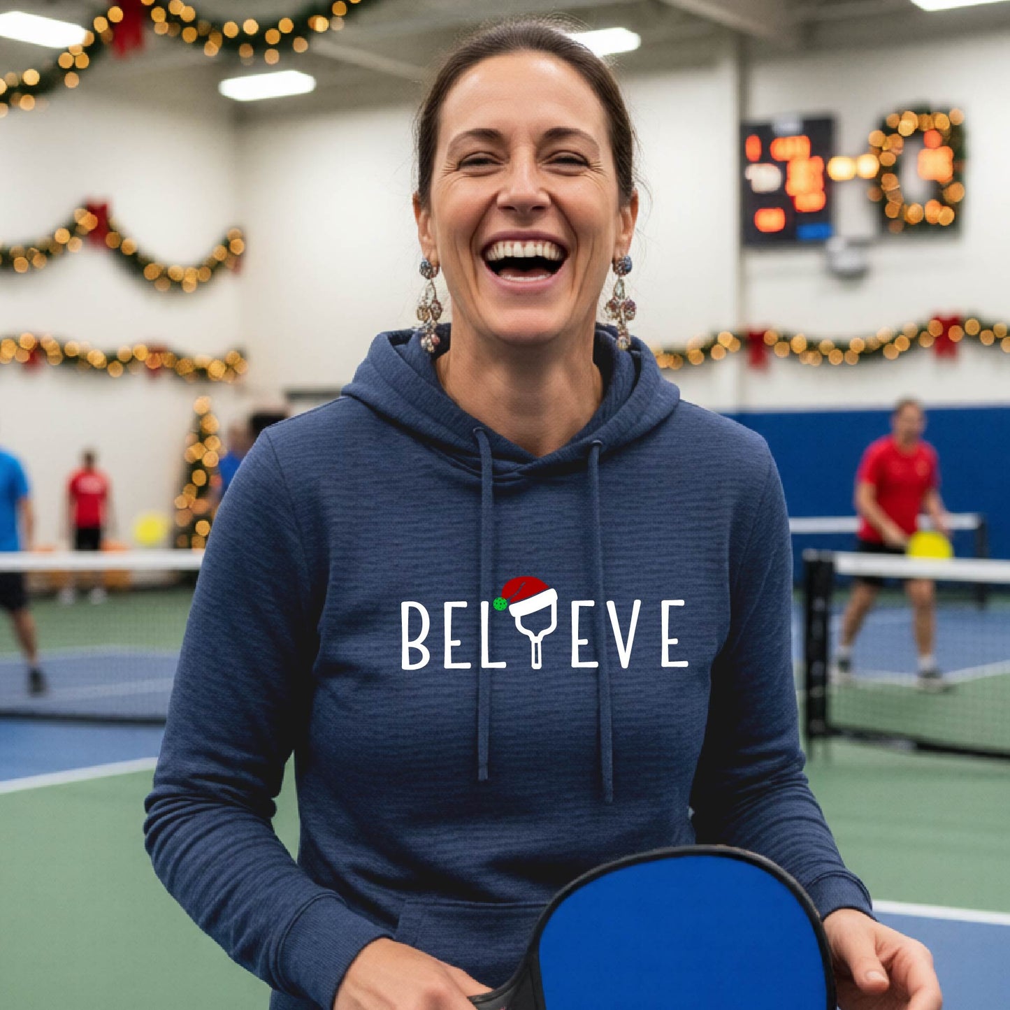 A navy blue hooded sweatshirt featuring a graphic print that reads "Believe" . The text I is replaced with a pickleball paddle wearing a santa hat.  The backgro0und is Christmas themed. 