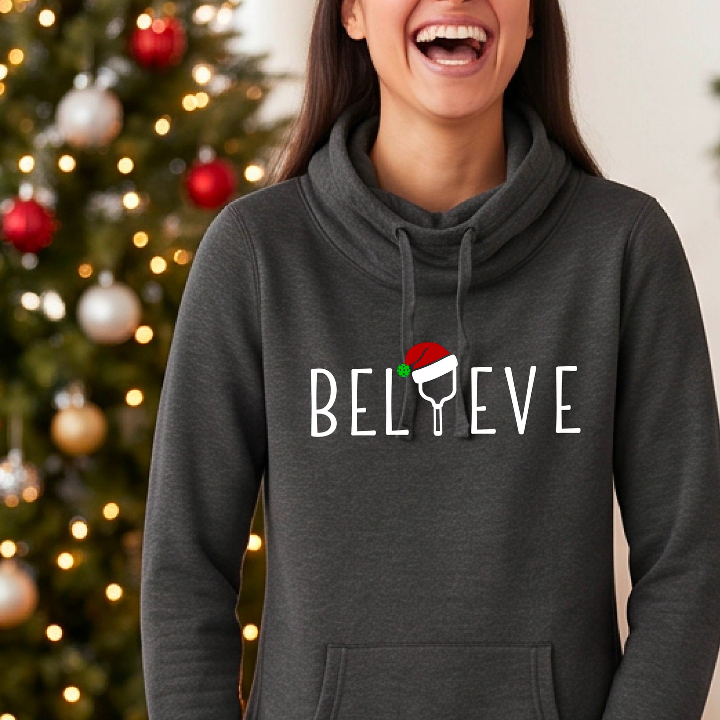 A heather black cowl-neck hooded sweatshirt featuring a graphic print that reads BGELIEVE" in white ext. The design includes a pickelball paddle wearing a santa hat.