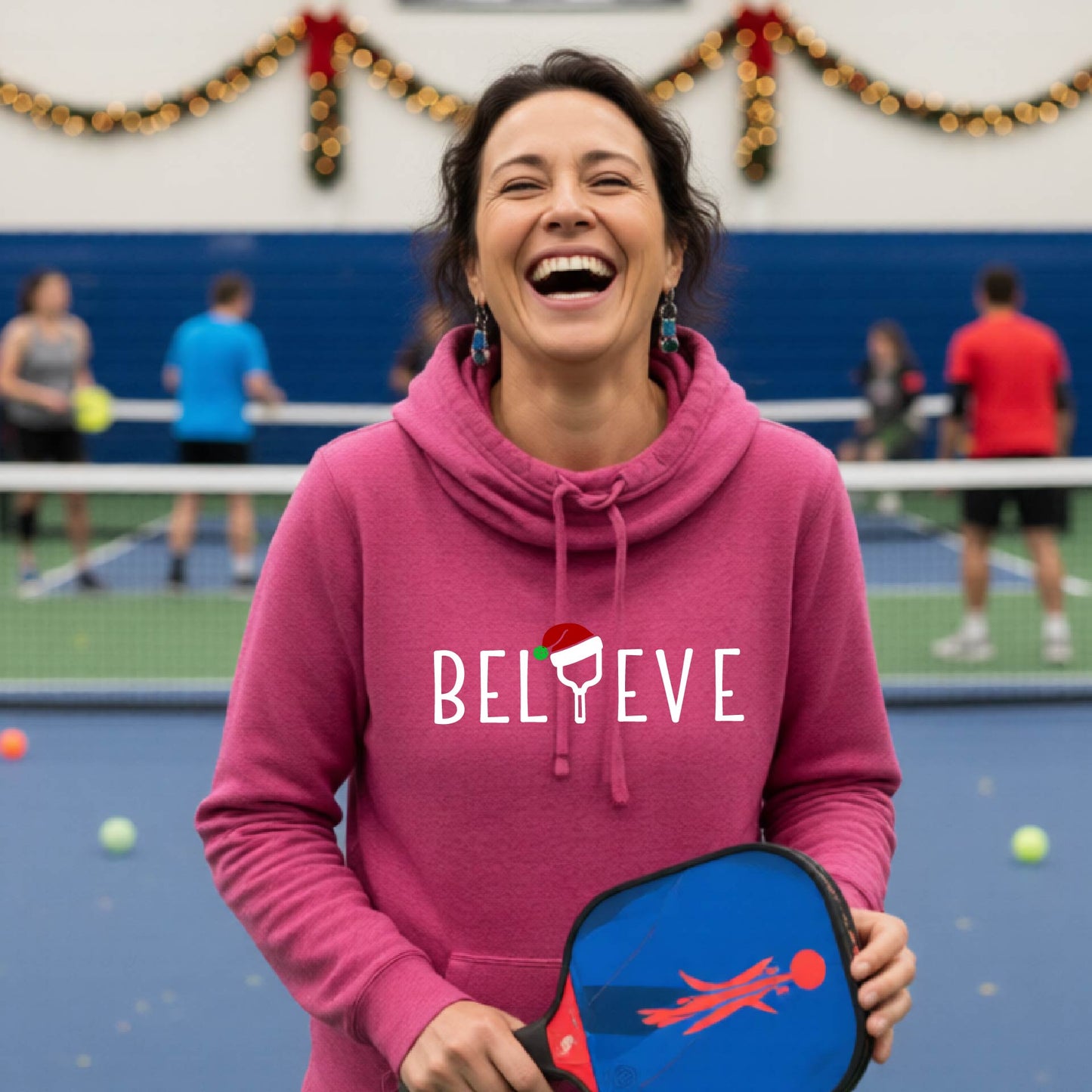 A pink cowl-neck hooded sweatshirt featuring a graphic print that reads BGELIEVE" in white ext. The design includes a pickelball paddle wearing a santa hat.