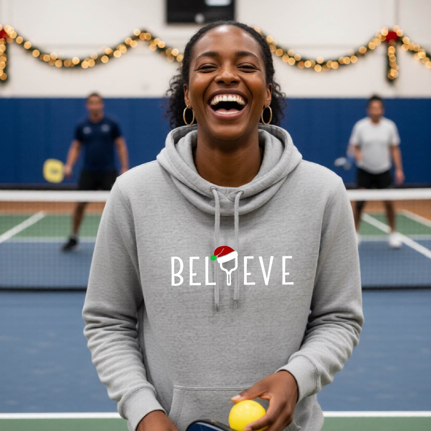 A gray cowl-neck hooded sweatshirt featuring a graphic print that reads BGELIEVE" in white ext. The design includes a pickelball paddle wearing a santa hat.