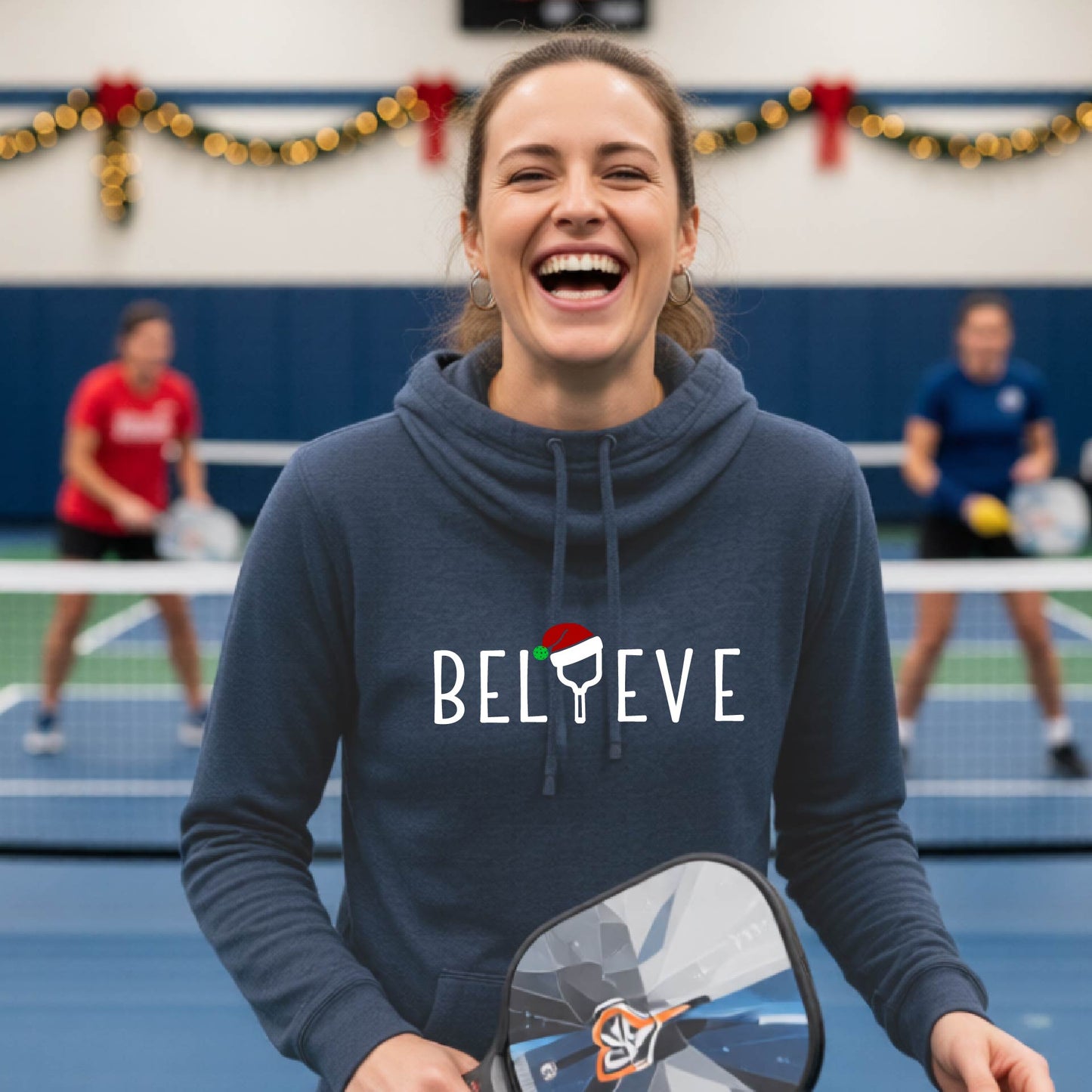 A navy blue cowl-neck hooded sweatshirt featuring a graphic print that reads BGELIEVE" in white ext. The design includes a pickelball paddle wearing a santa hat.