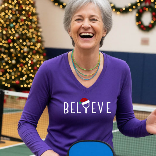 A women's purple long-sleeve v-neck shirt featuring a pickleball graphic with text "BELIEVE"  with the I replaced by a pickleball paddle wearing a santa hat and  Christmas themed background.themed backgrou dat 