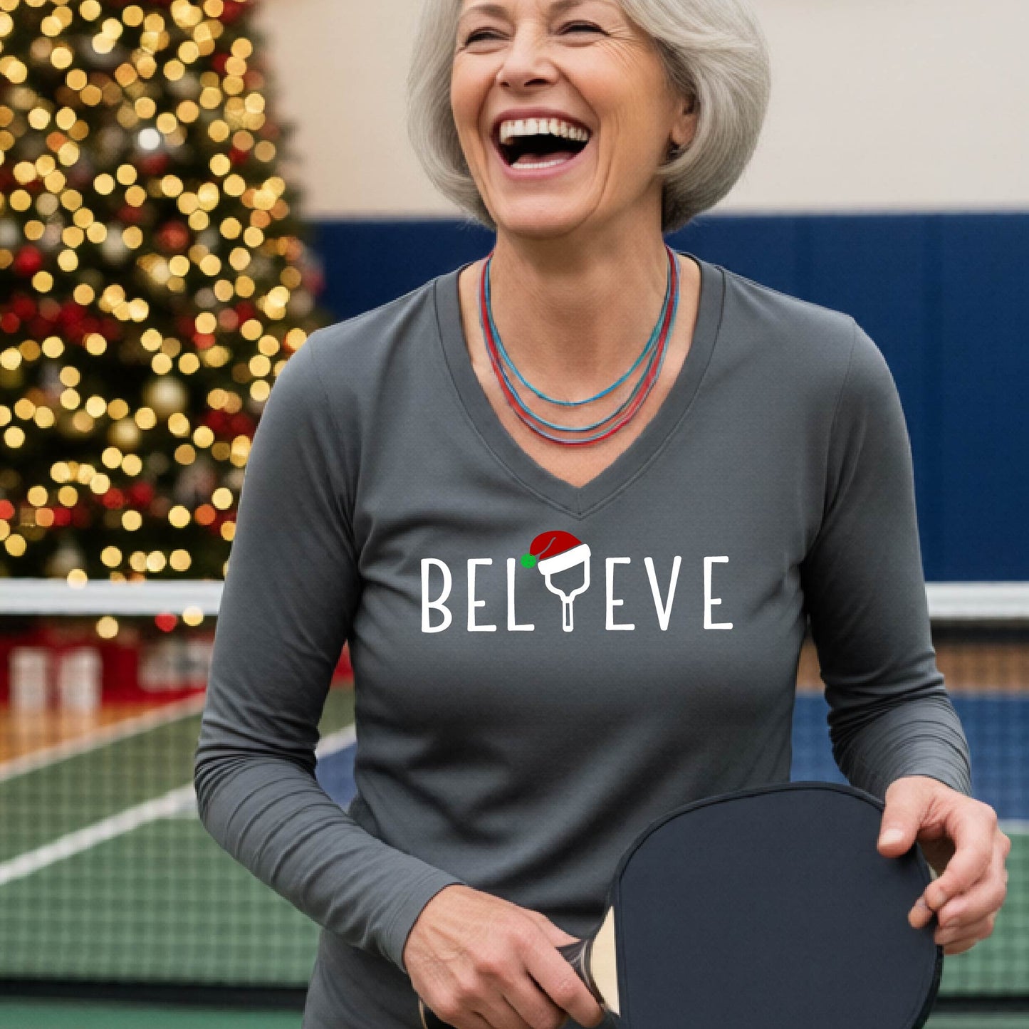 A women's grey long-sleeve v-neck shirt featuring a pickleball graphic with text "BELIEVE"  with the I replaced by a pickleball paddle wearing a santa hat and  Christmas themed background.