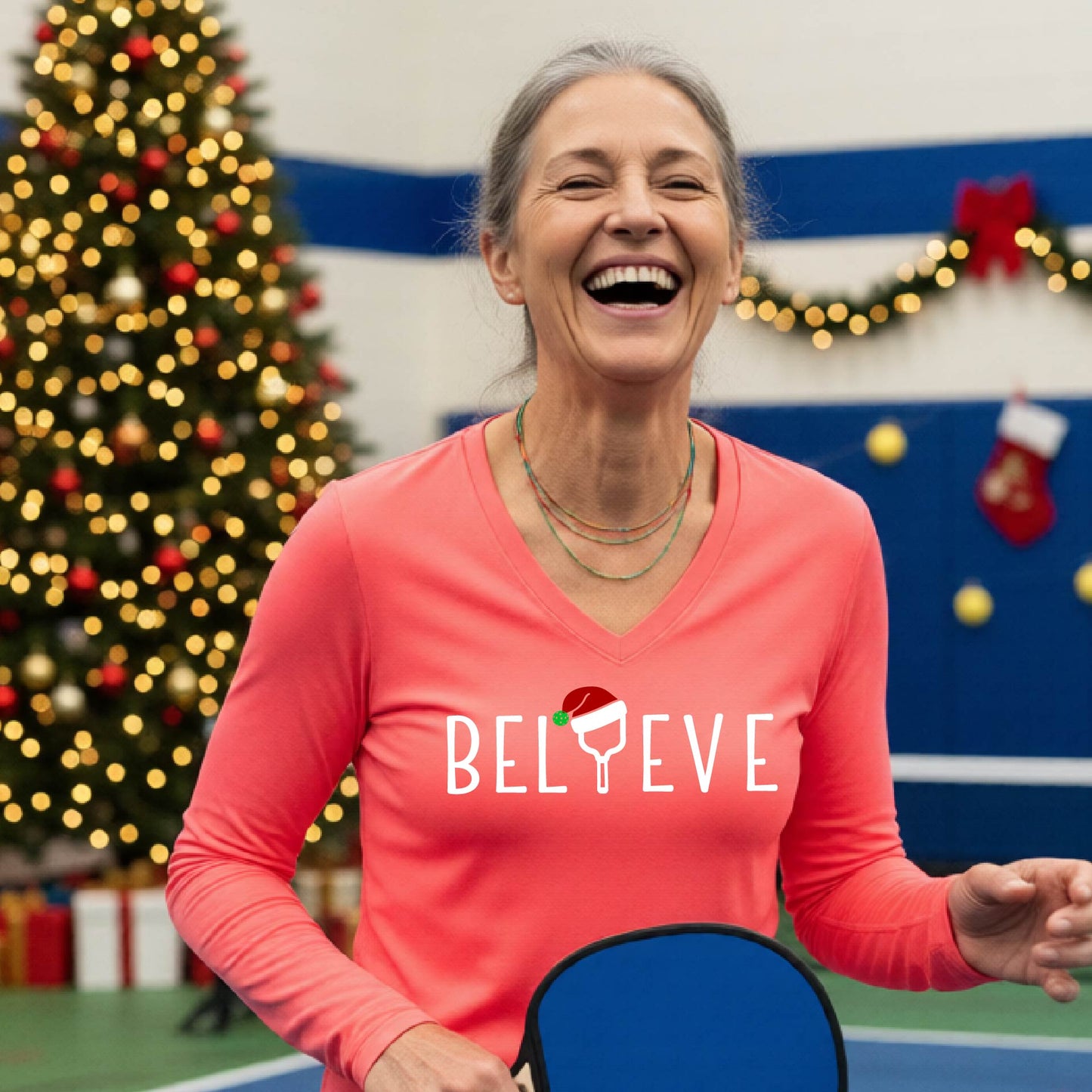A women's coral pink long-sleeve v-neck shirt featuring a pickleball graphic with text "BELIEVE"  with the I replaced by a pickleball paddle wearing a santa hat and  Christmas themed background.