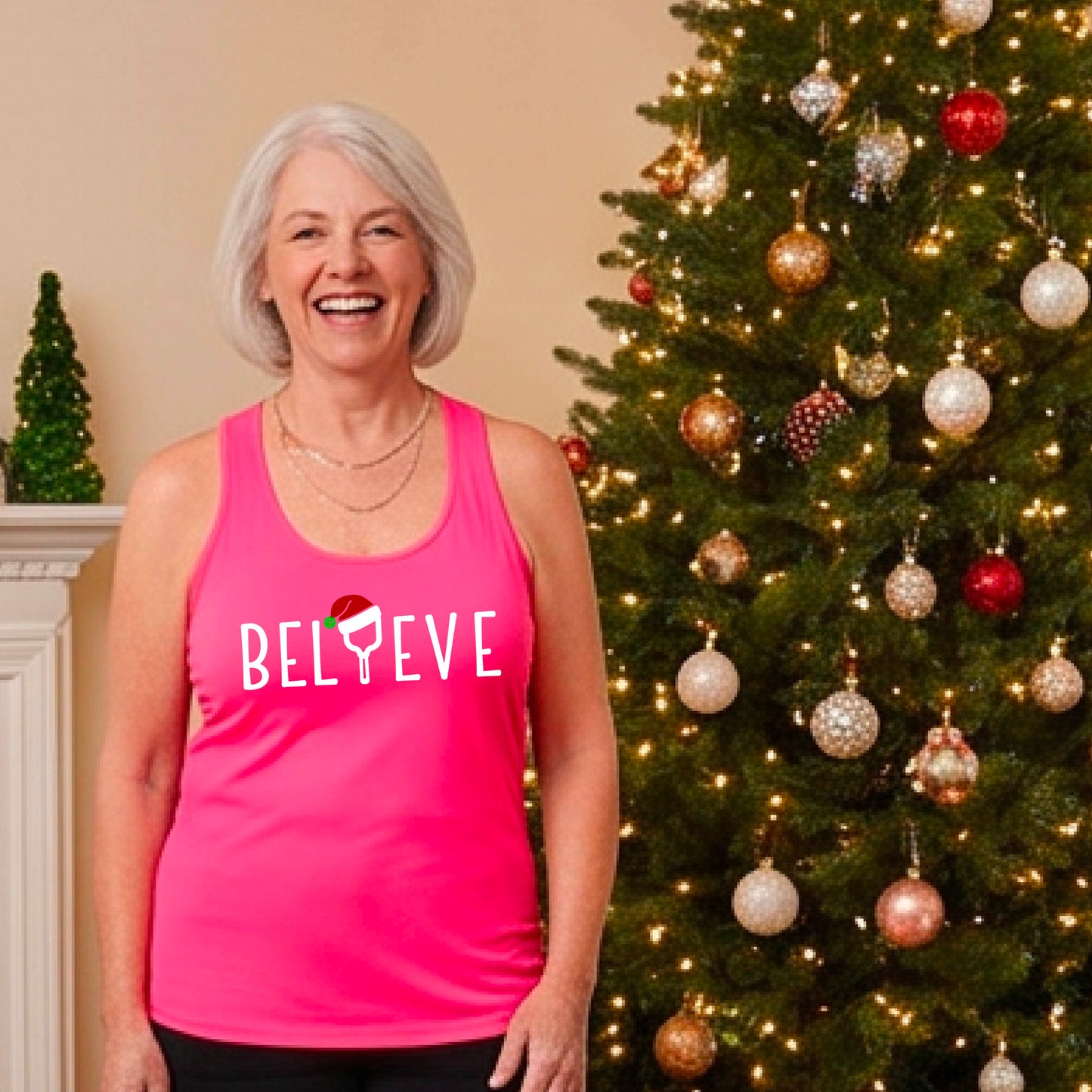 A smiling woman wears a bright pink racerback tank top with the word 'BELIEVE' in white, where the letter 'I' is replaced by a pickleball paddle wearing a Santa hat. She stands in front of a lit Christmas tree with ornaments.