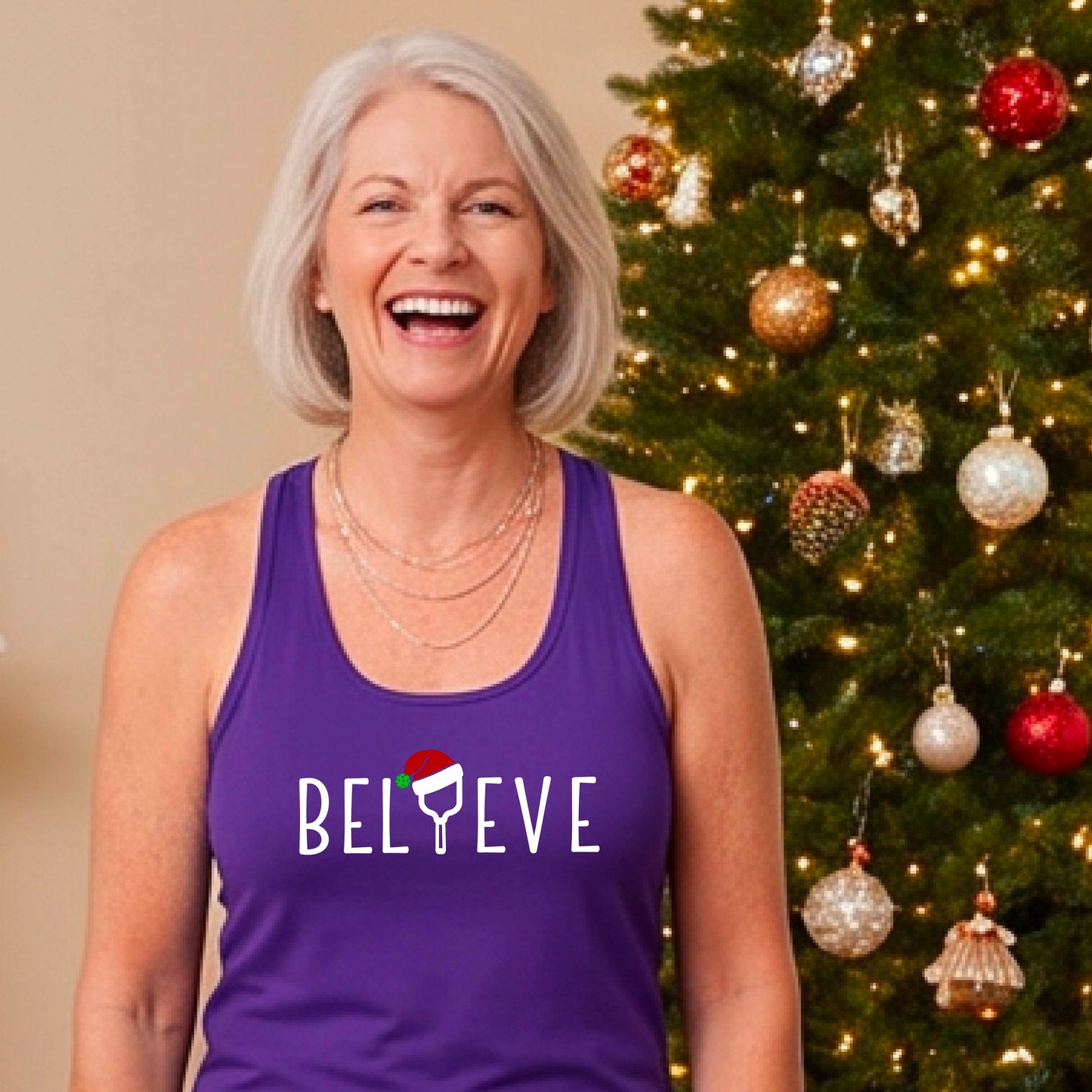A smiling woman wears a purple racerback tank top with the word 'BELIEVE' in white, where the letter 'I' is replaced by a pickleball paddle wearing a Santa hat. She stands in front of a lit Christmas tree with ornaments.