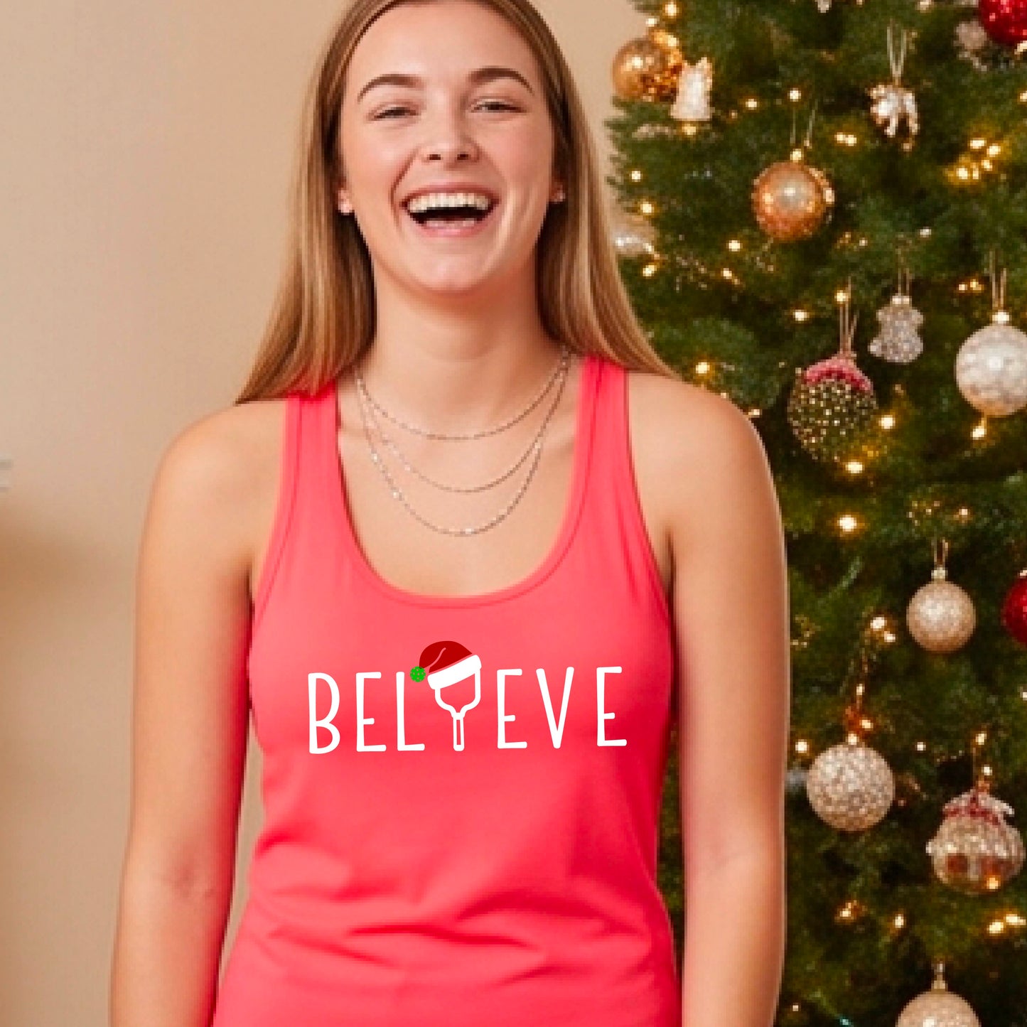 A smiling woman wears a coral pink racerback tank top with the word 'BELIEVE' in white, where the letter 'I' is replaced by a pickleball paddle wearing a Santa hat. She stands in front of a lit Christmas tree with ornaments.
