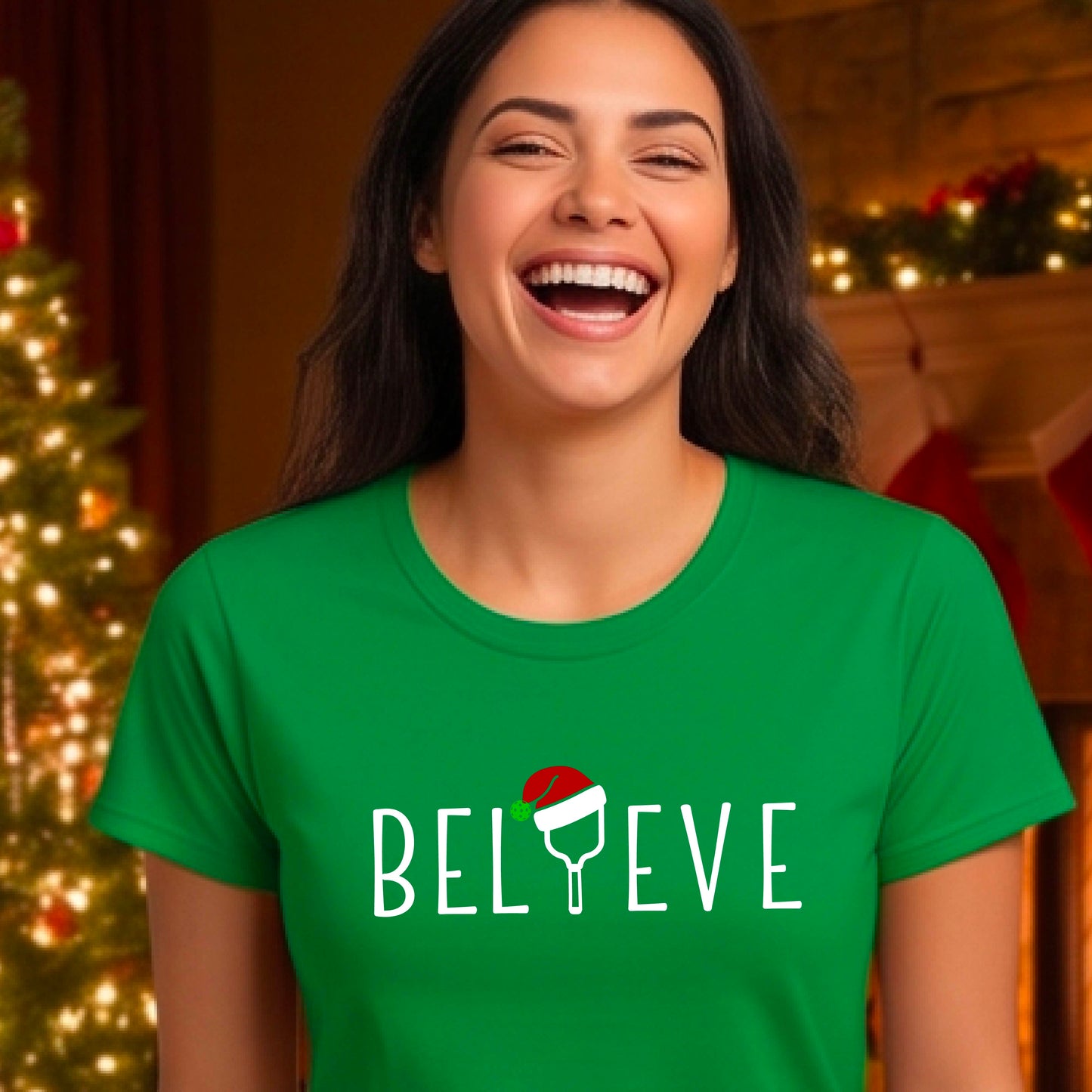 A woman wearing a green graphic t-shirt with the word 'BELIEVE' in white, where the letter 'I' is replaced by a pickleball topped with a red Santa hat. The background shows a festive holiday scene with a decorated Christmas tree and warm string lights.