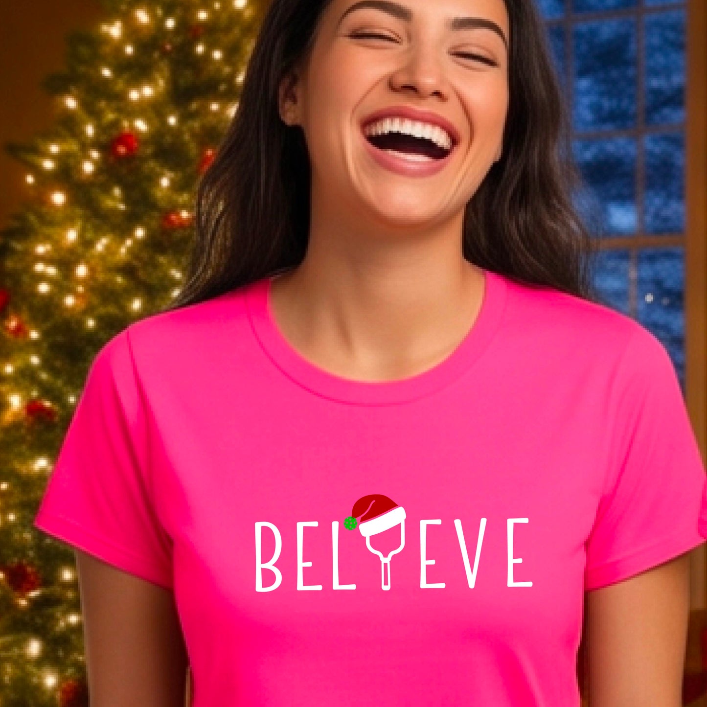 A woman wearing a bright pink graphic t-shirt with the word 'BELIEVE' in white, where the letter 'I' is replaced by a pickleball topped with a red Santa hat. The background shows a festive holiday scene with a decorated Christmas tree and warm string lights.