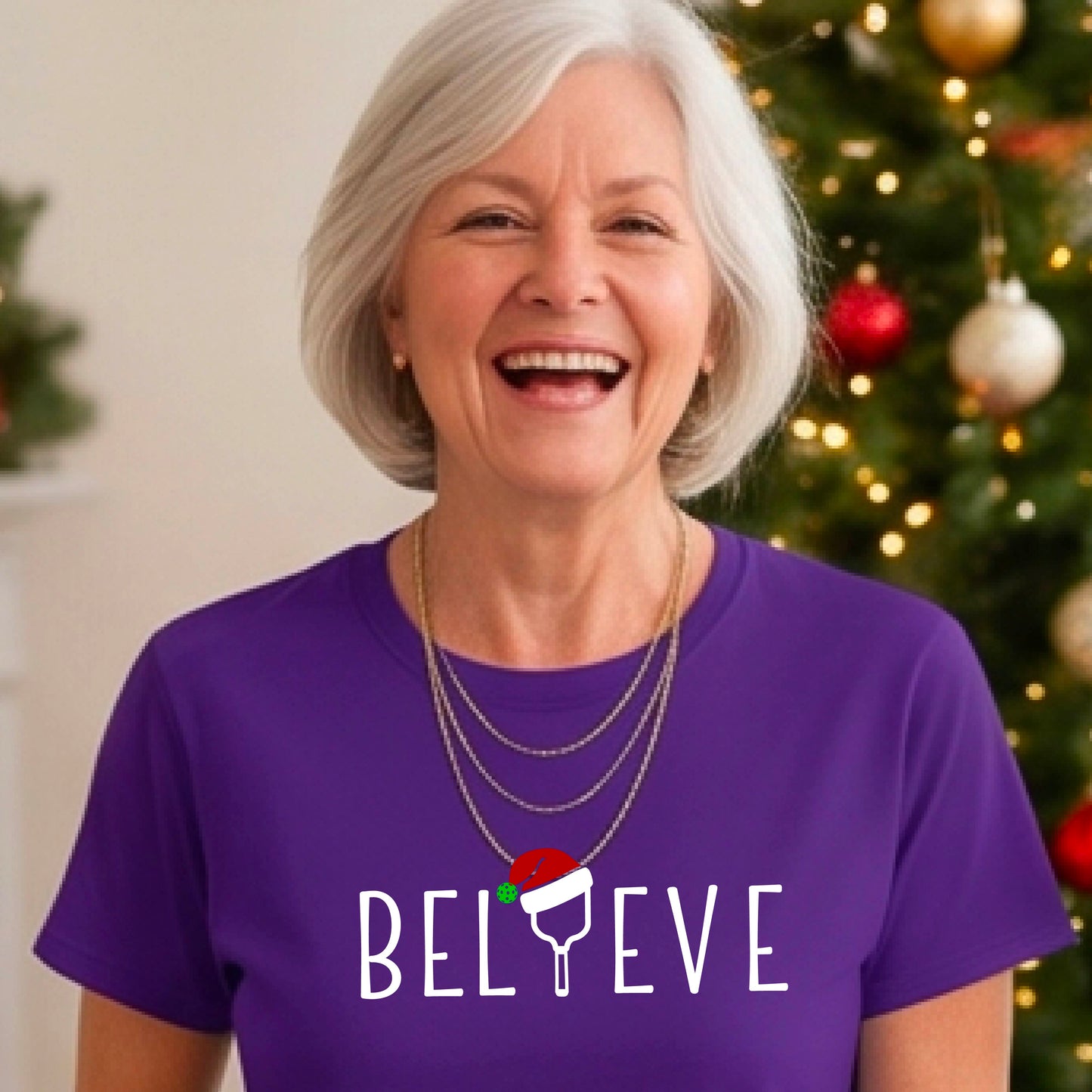 A woman wearing a purple graphic t-shirt with the word 'BELIEVE' in white, where the letter 'I' is replaced by a pickleball topped with a red Santa hat. The background shows a festive holiday scene with a decorated Christmas tree and warm string lights.
