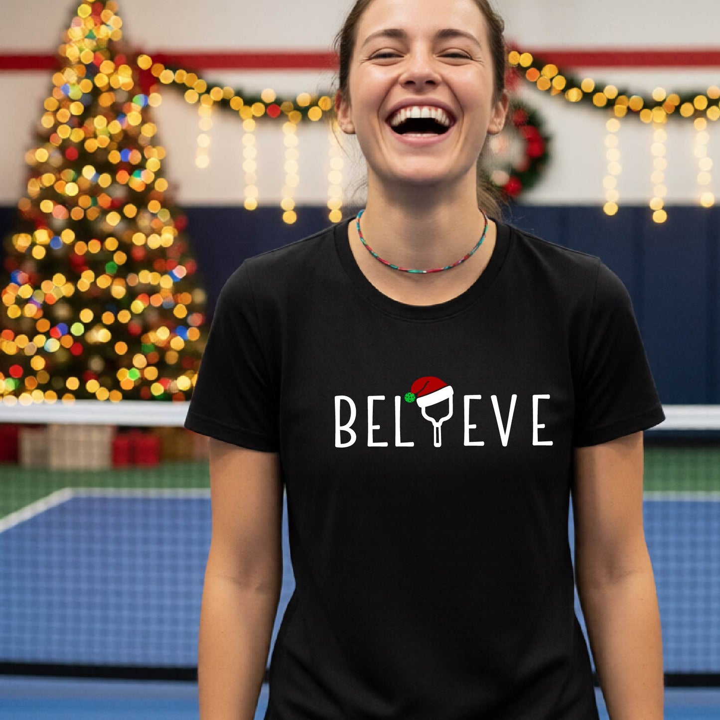 A woman wearing a black graphic t-shirt with the word 'BELIEVE' in white, where the letter 'I' is replaced by a pickleball topped with a red Santa hat. The background shows a festive holiday scene with a decorated Christmas tree and warm string lights.