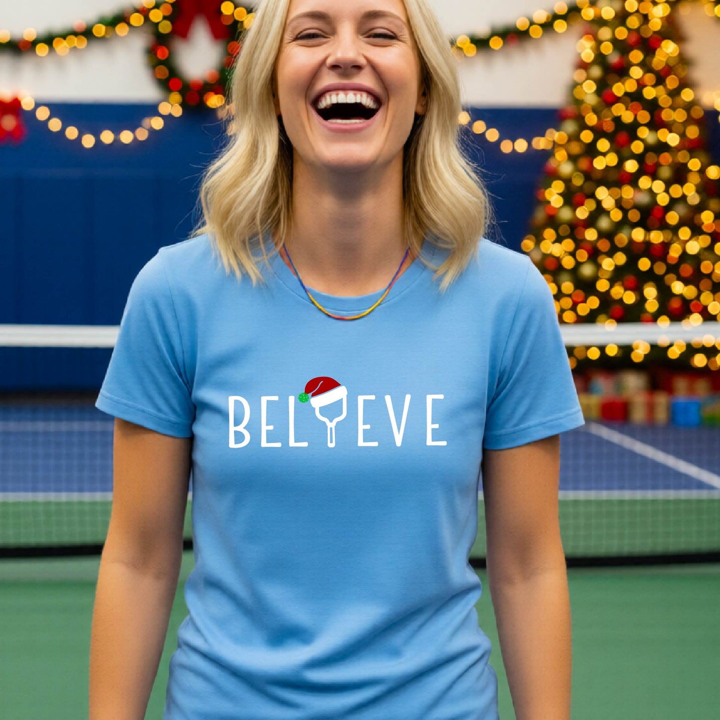 A woman wearing a light blue graphic t-shirt with the word 'BELIEVE' in white, where the letter 'I' is replaced by a pickleball topped with a red Santa hat. The background shows a festive holiday scene with a decorated Christmas tree and warm string lights.