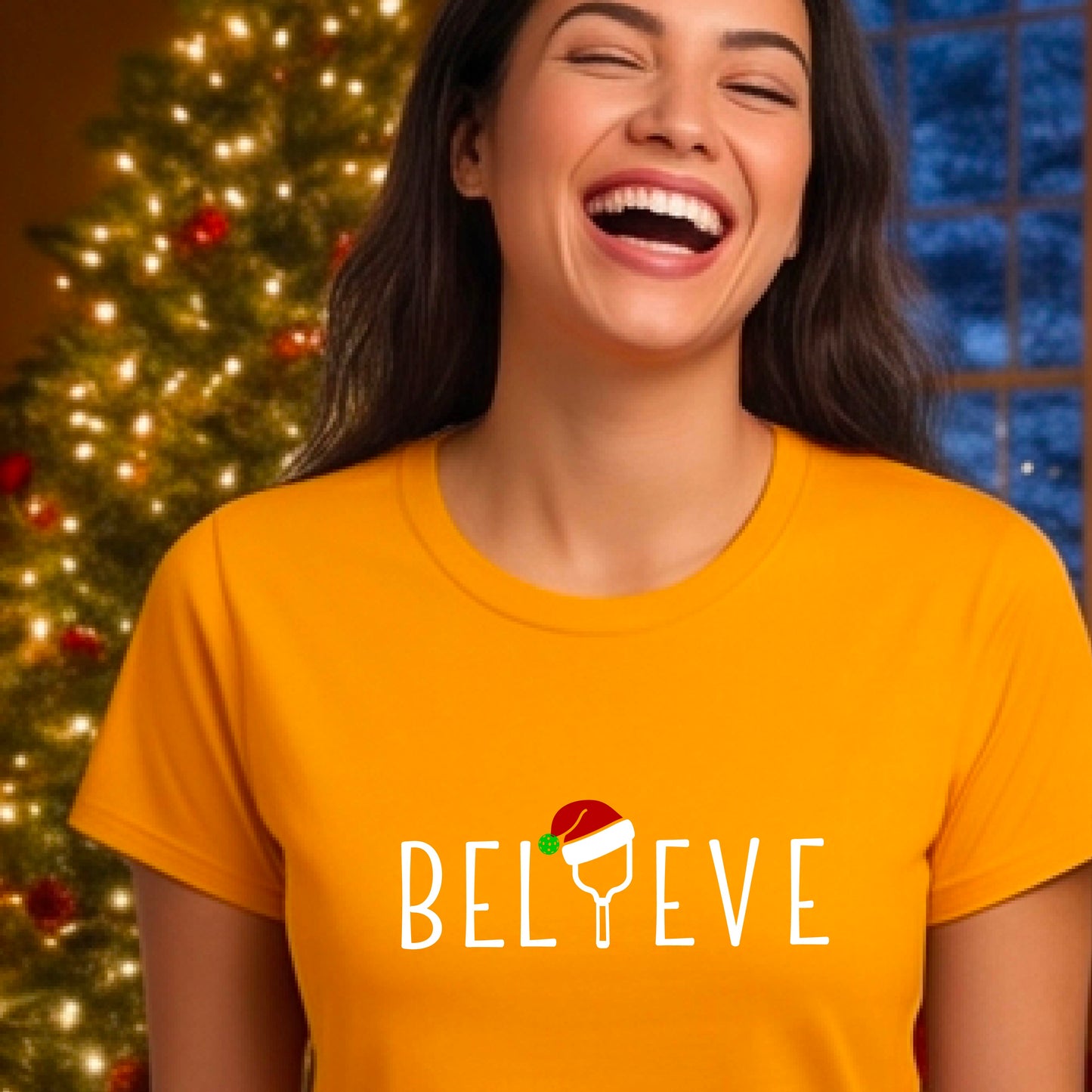 A woman wearing a mustard yellow gold graphic t-shirt with the word 'BELIEVE' in white, where the letter 'I' is replaced by a pickleball topped with a red Santa hat. The background shows a festive holiday scene with a decorated Christmas tree and warm string lights.