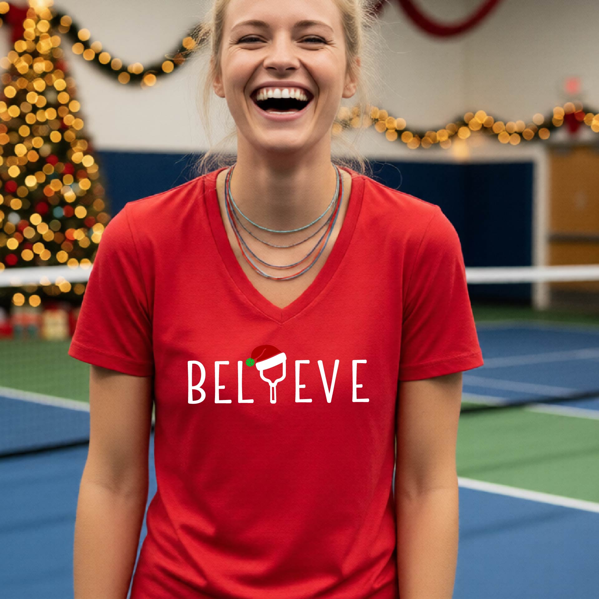 A woman smiling on an indoor pickleball court wearing a red V-neck t-shirt with the word 'BELIEVE' in white, where the letter 'I' is replaced by a pickleball paddle  graphic wearing a red and green Santa hat.