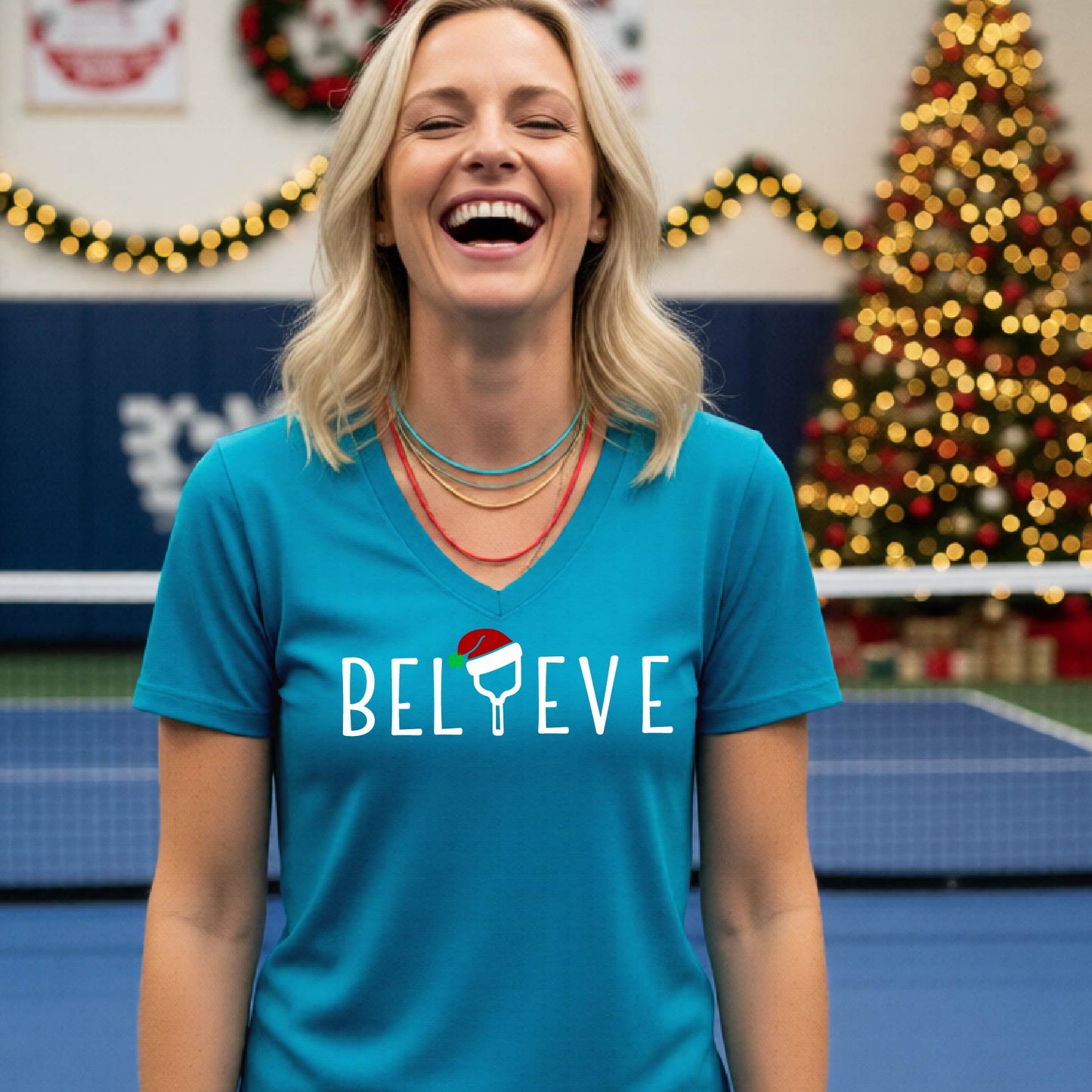 A woman smiling on an indoor pickleball court wearing a teal V-neck t-shirt with the word 'BELIEVE' in white, where the letter 'I' is replaced by a pickleball paddle  graphic wearing a red and green Santa hat.