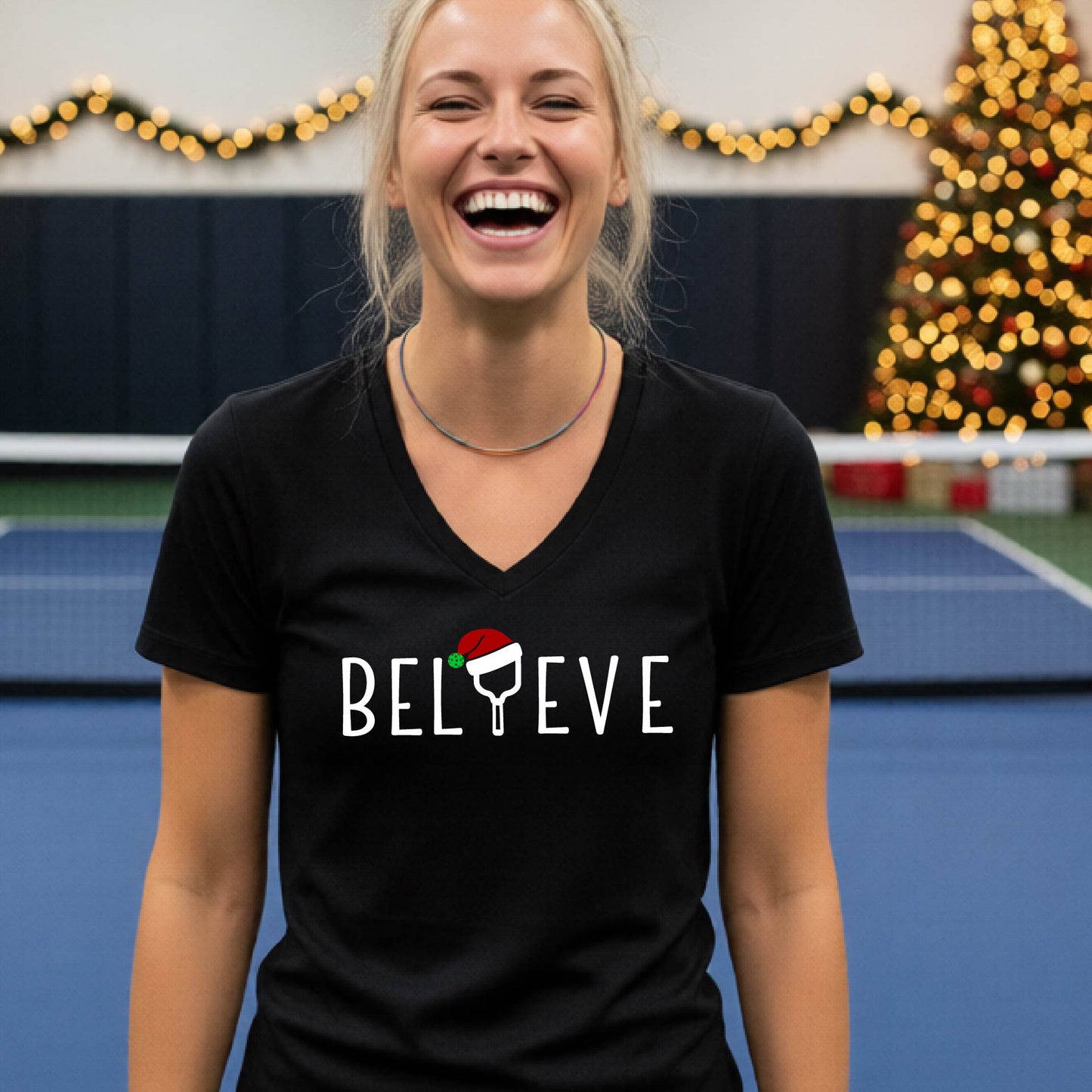 A woman smiling on an indoor pickleball court wearing a black V-neck t-shirt with the word 'BELIEVE' in white, where the letter 'I' is replaced by a pickleball paddle  graphic wearing a red and green Santa hat.