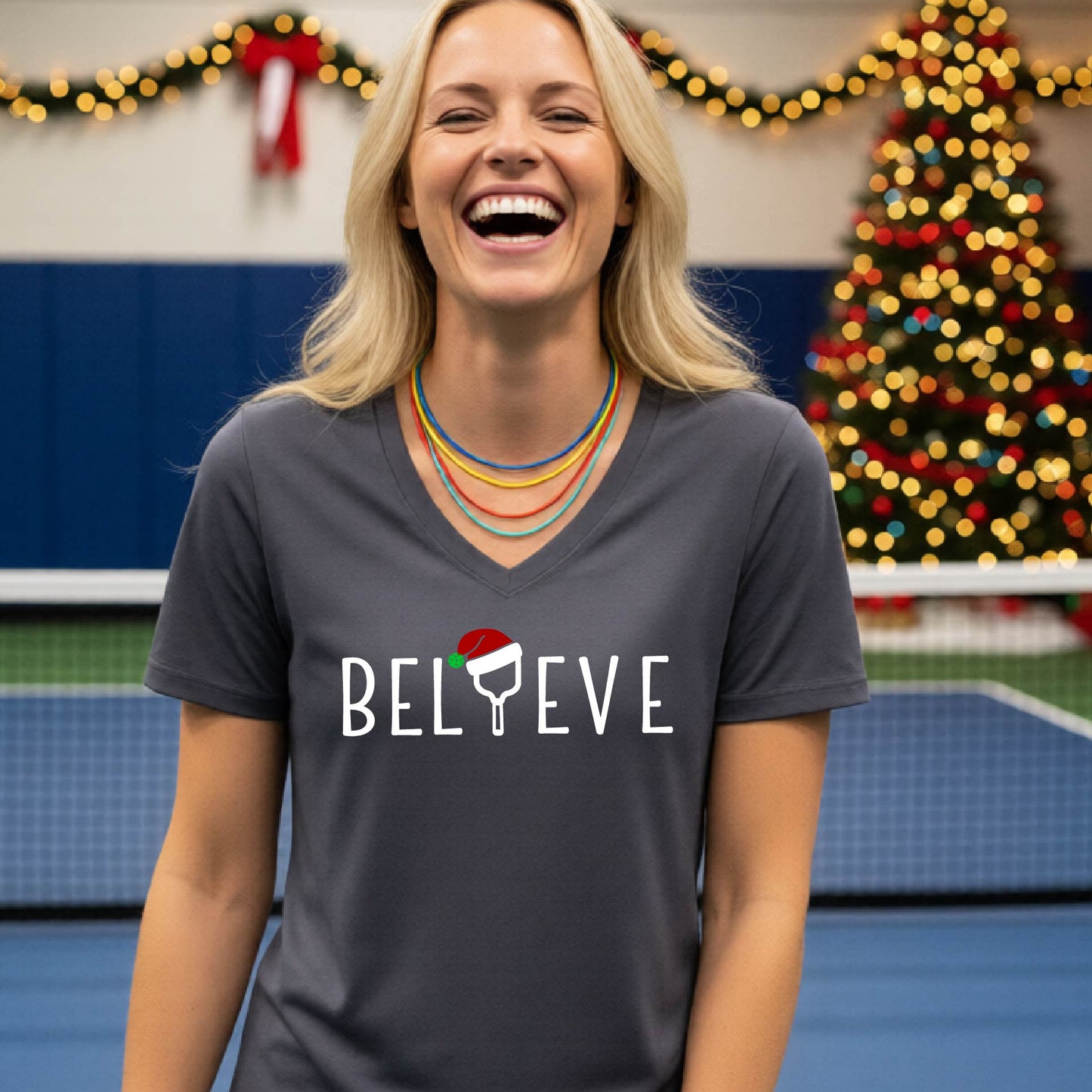 A woman smiling on an indoor pickleball court wearing a dark gray V-neck t-shirt with the word 'BELIEVE' in white, where the letter 'I' is replaced by a pickleball paddle  graphic wearing a red and green Santa hat.