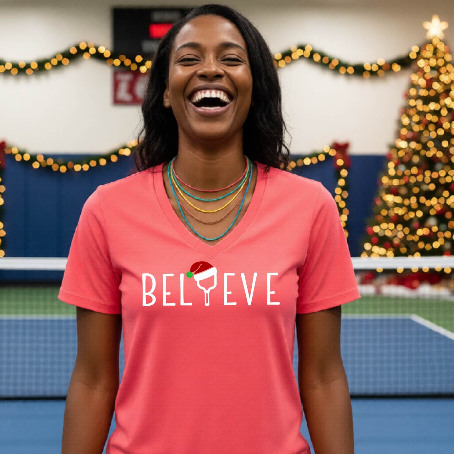 A woman smiling on an indoor pickleball court wearing a coral-pink V-neck t-shirt with the word 'BELIEVE' in white, where the letter 'I' is replaced by a pickleball paddle  graphic wearing a red and green Santa hat.