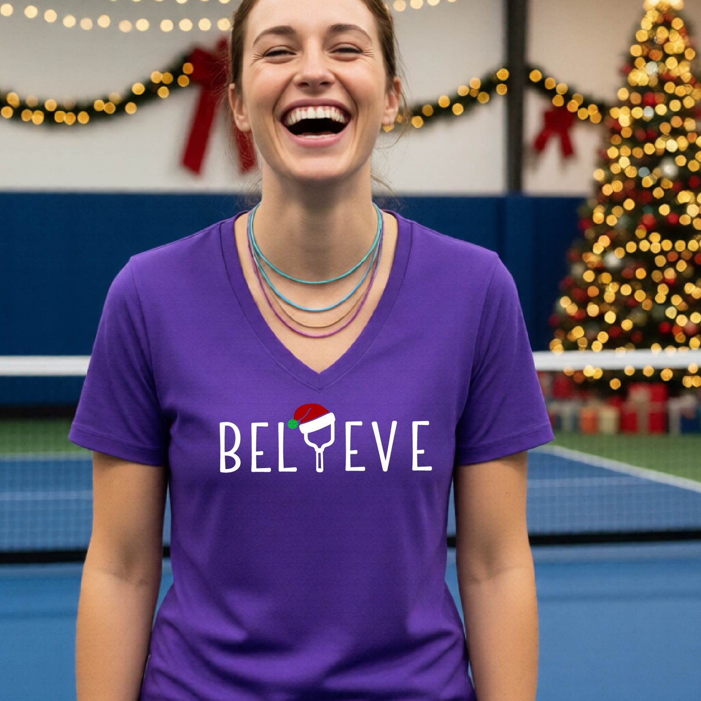 A woman smiling on an indoor pickleball court wearing a purple V-neck t-shirt with the word 'BELIEVE' in white, where the letter 'I' is replaced by a pickleball paddle  graphic wearing a red and green Santa hat.