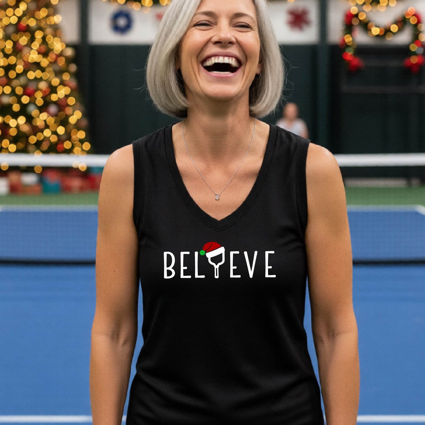 A smiling woman stands on a blue pickleball court wearing a black sleeveless tank top with the word 'BELIEVE' in white, where the 'I' is replaced by a pickleball paddle  wearing a red Santa hat.