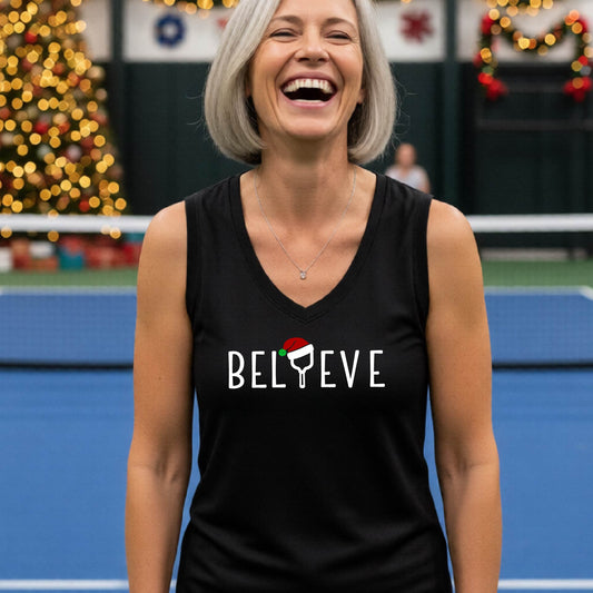 A smiling woman stands on a blue pickleball court wearing a black sleeveless tank top with the word 'BELIEVE' in white, where the 'I' is replaced by a pickleball paddle  wearing a red Santa hat.