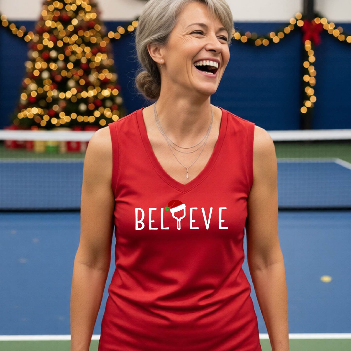A smiling woman stands on a blue pickleball court wearing a red sleeveless tank top with the word 'BELIEVE' in white, where the 'I' is replaced by a pickleball paddle  wearing a red Santa hat.