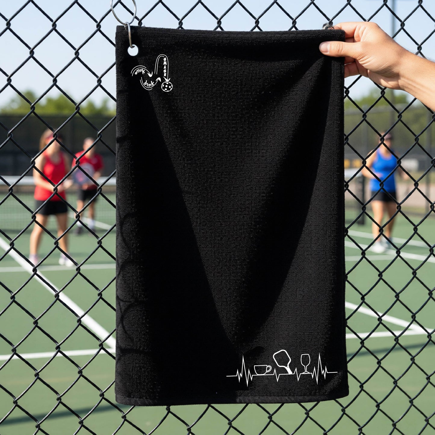 A person holds a black court towel grommet with a pickleball court blurred in the background. The towel has a white line-art graphic of a coffee cup, pickleball paddle and a wine glass, on a heartbeat line on the bottom right with a DINK DINK SMASH logo in the top left corner.