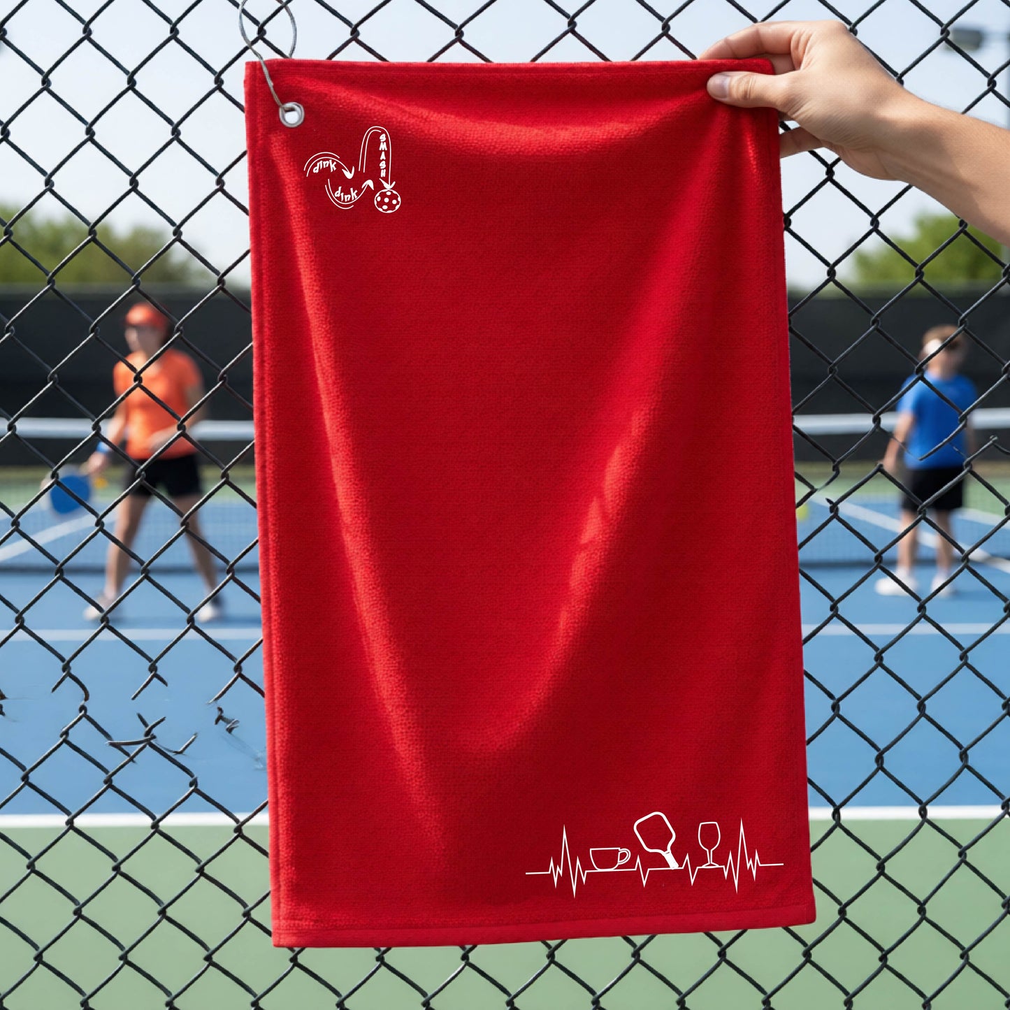 A person holds a red court towel grommet with a pickleball court blurred in the background. The towel has a white line-art graphic of a coffee cup, pickleball paddle and a wine glass, on a heartbeat line on the bottom right with a DINK DINK SMASH logo in the top left corner.