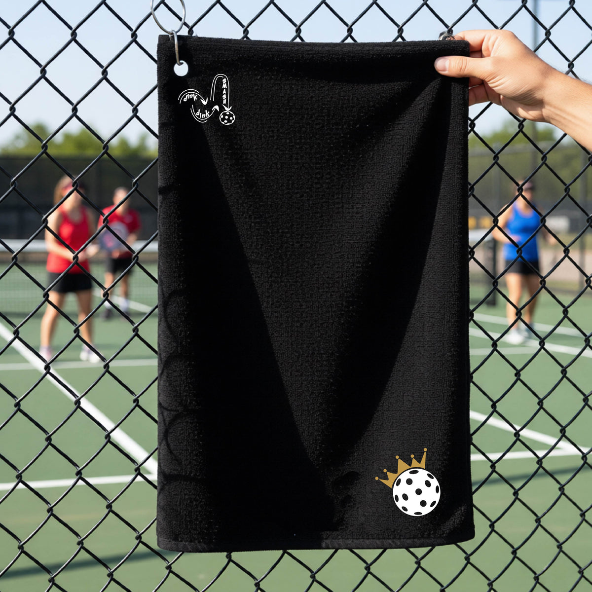 A close up of a black grommet and hook pickleball court towel with a DINK DINK SMASH graphic in the upper left corner and a white pickleball crown with ball graphic in the bottom right corner. 