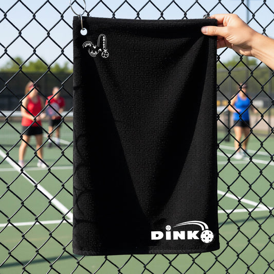 A close up of a black pickleball court towel with a log of 'DINK DINK SMASH' in the upper left corner and a pickleball design of text 'DINK' with a white pickleall in flight, with a pickleball court in the background. 