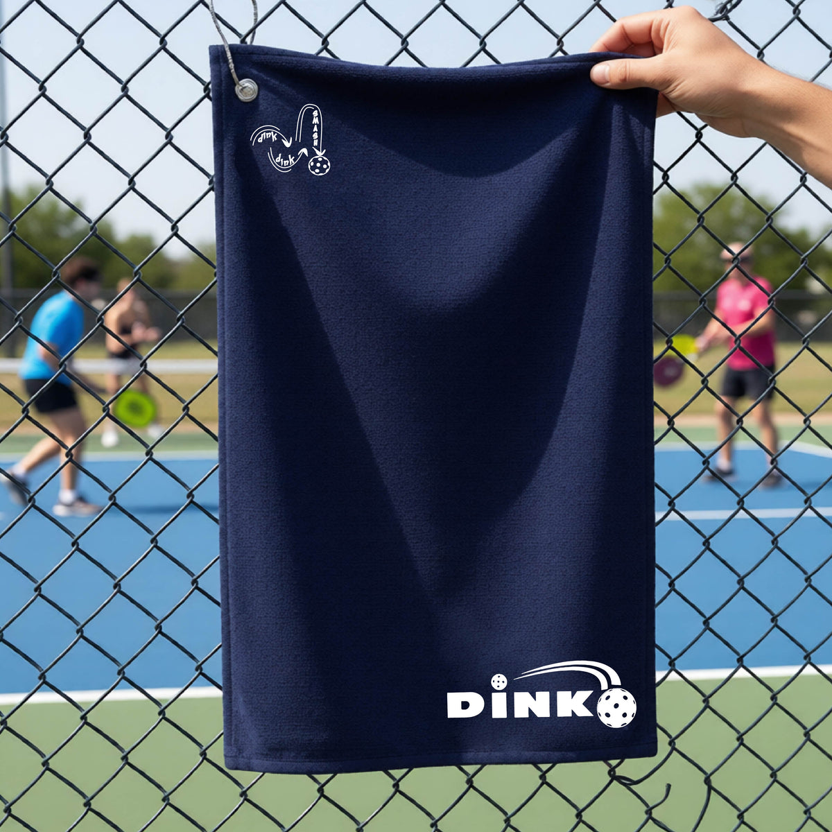 A close up of a navy blue pickleball court towel with a log of 'DINK DINK SMASH' in the upper left corner and a pickleball design of text 'DINK' with a white pickleall in flight, with a pickleball court in the background. 